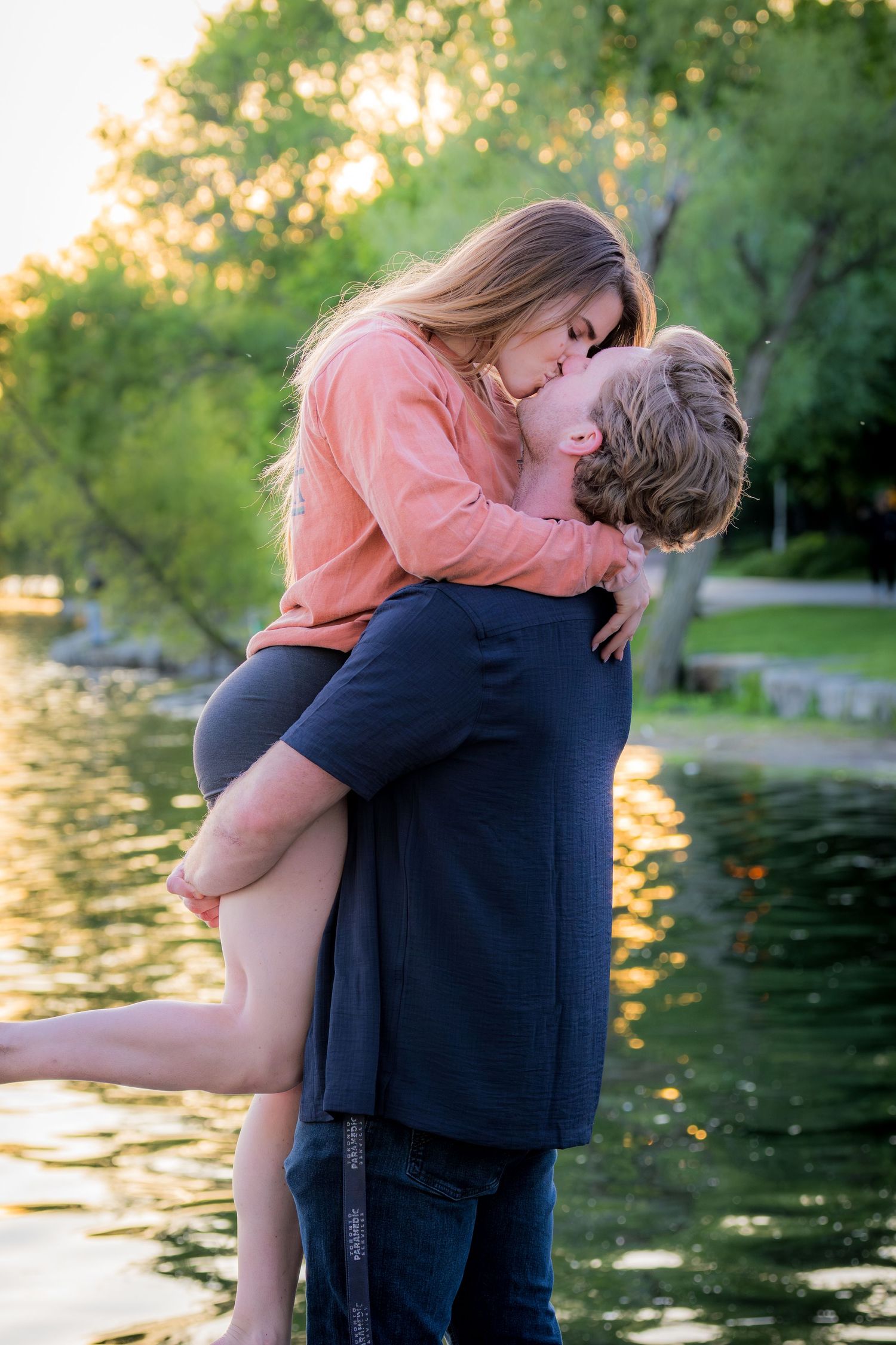 Man lifting woman for a kiss near a pond surrounded by green trees at golden hour.
