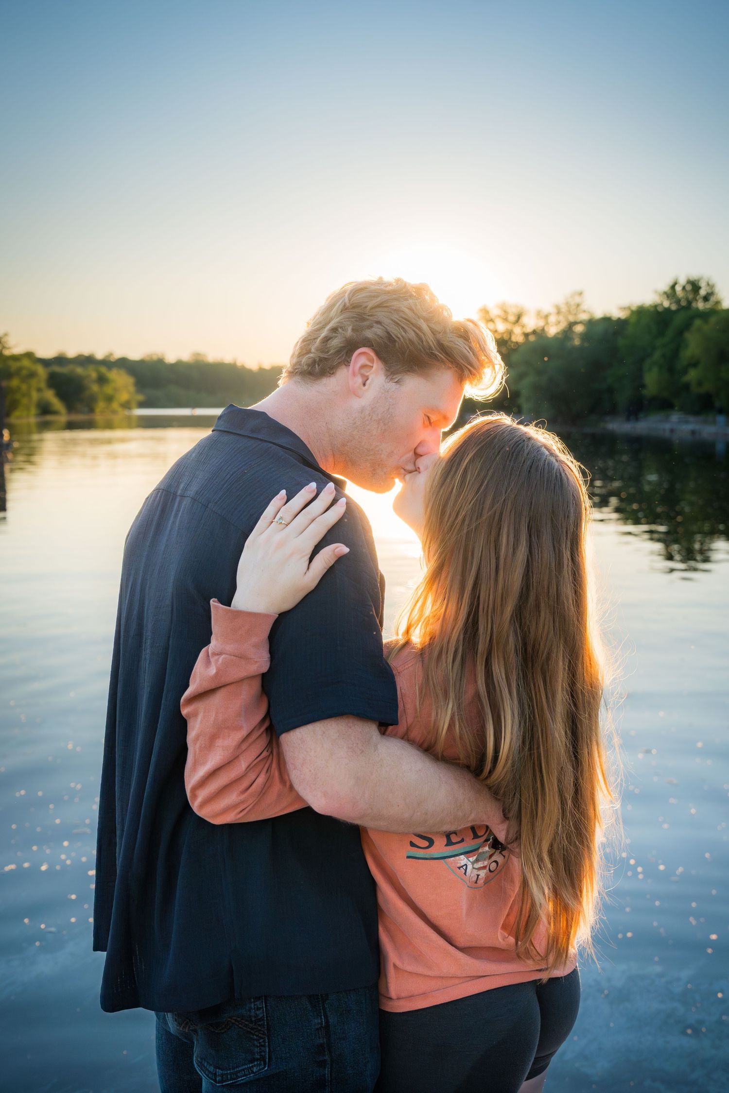 Couple sharing a tender kiss at golden hour by a serene lake, with warm sunset light glowing behind them.