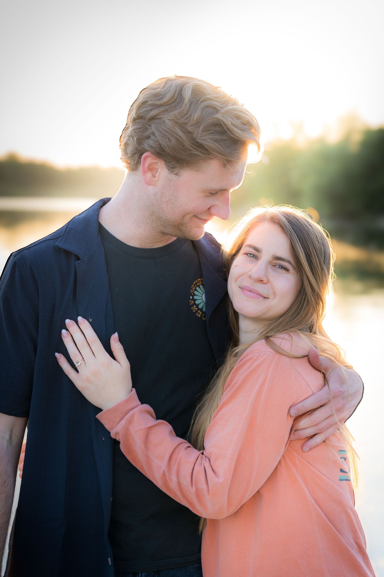 Newly engaged couple embracing near a lake at sunset, woman showing her engagement ring.