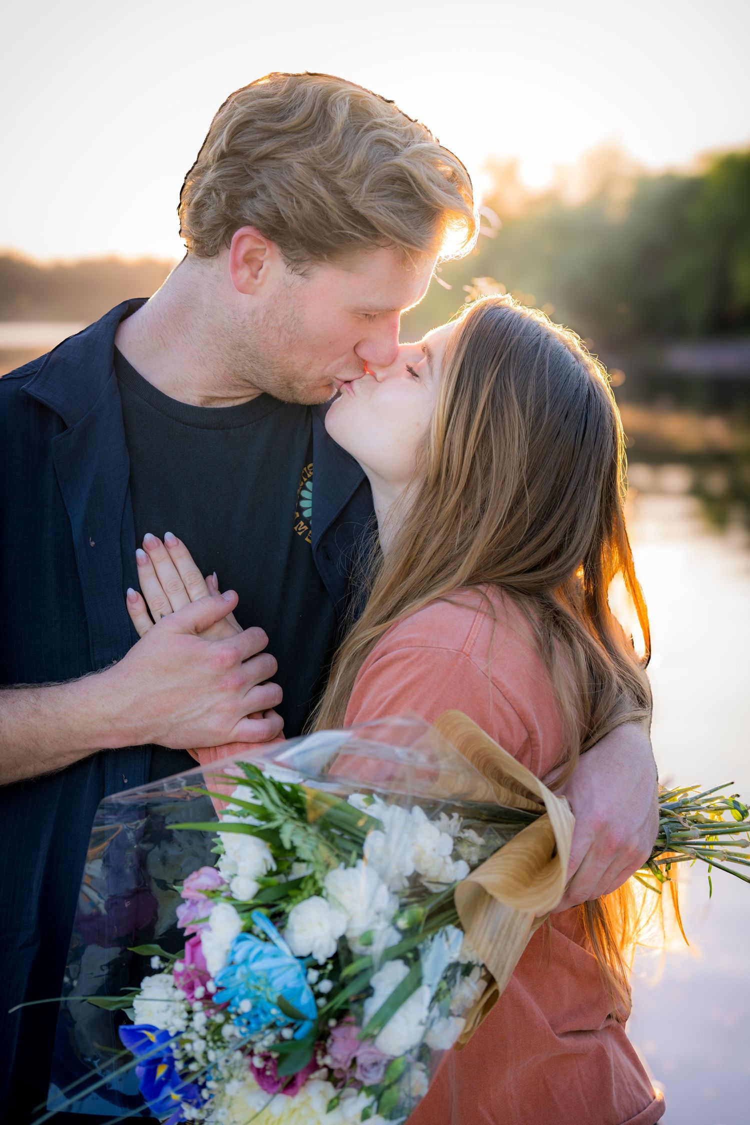Couple sharing a passionate kiss while holding a bouquet of flowers by a lake at sunset.