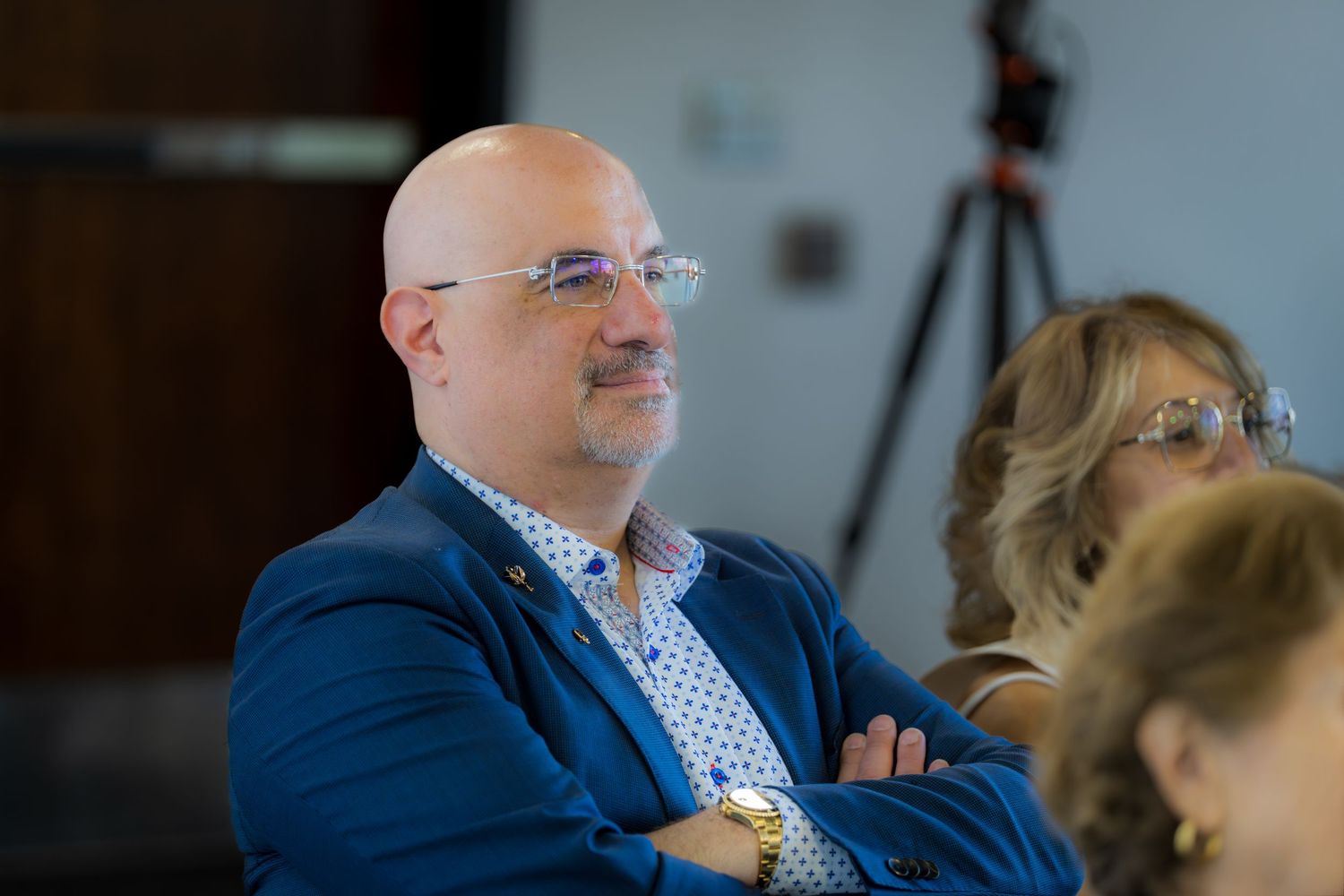 Bald man with glasses and blue blazer smiles with arms crossed at formal event.