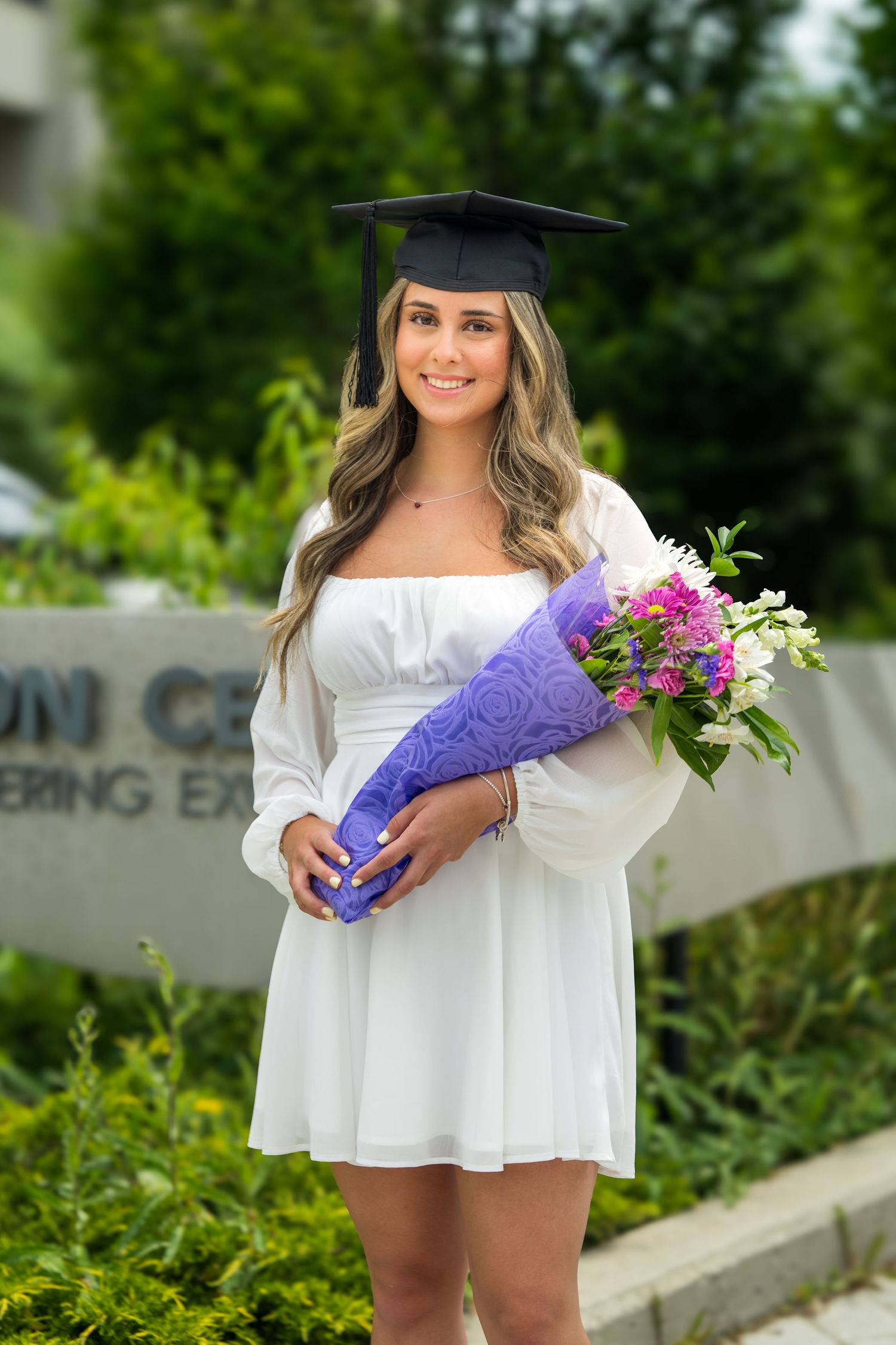 Graduate in flowing white dress holds purple bouquet during outdoor photo session.