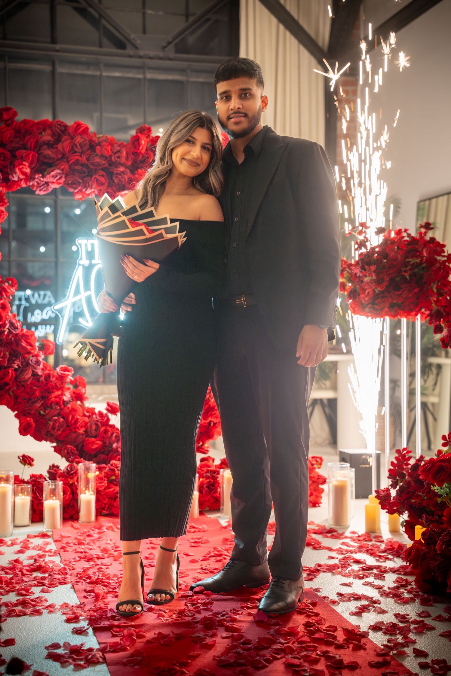 Couple posing in romantic setting with sparklers, red roses, and neon lighting.