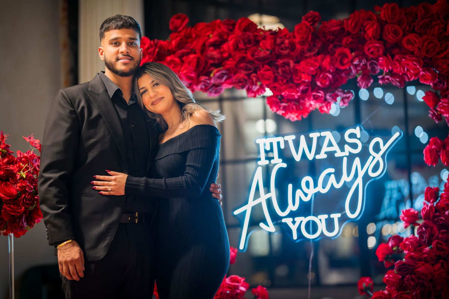 Romantic pair embraces by illuminated sign and red rose wall in evening formal wear.