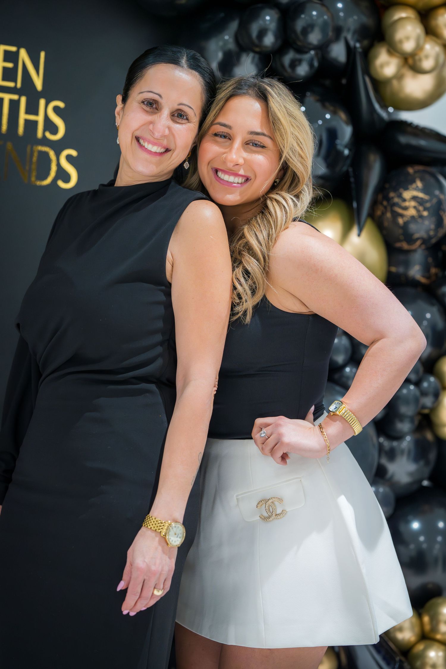 Two people in black dresses pose against a backdrop of black and gold balloons.