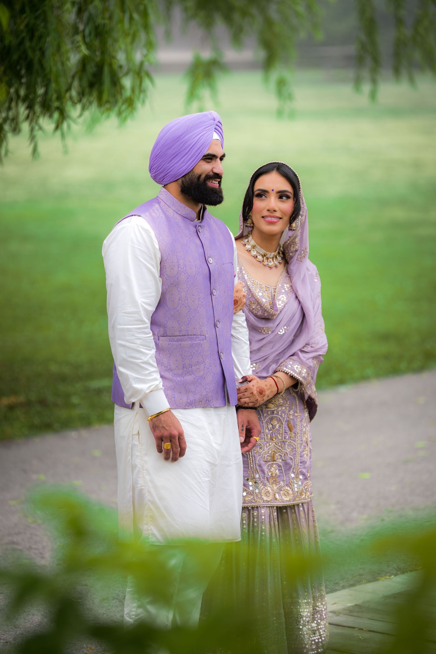A couple in coordinated purple and white traditional Punjabi outfits poses together in a garden setting.