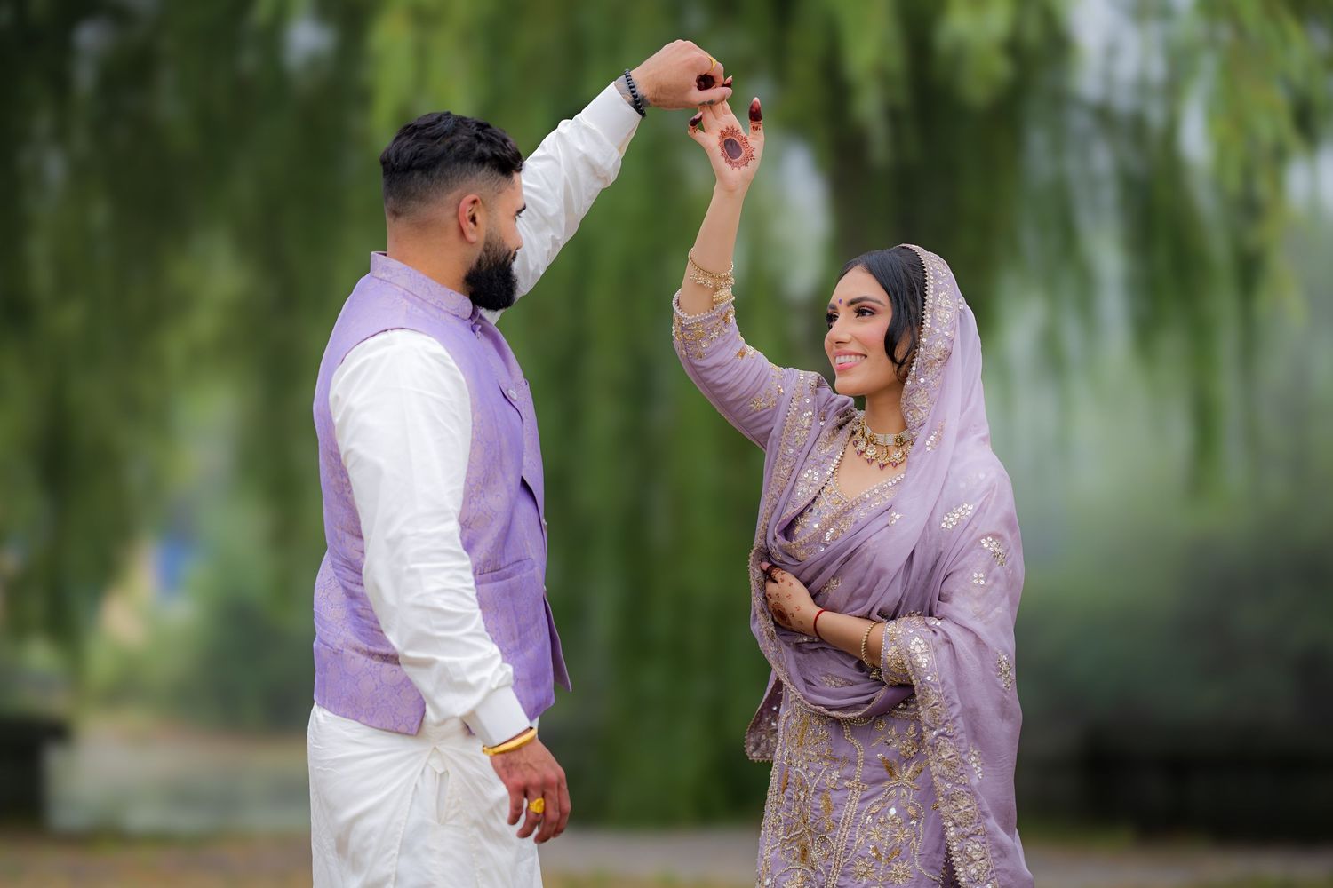 A couple in purple and white traditional dress dance together under willow branches.