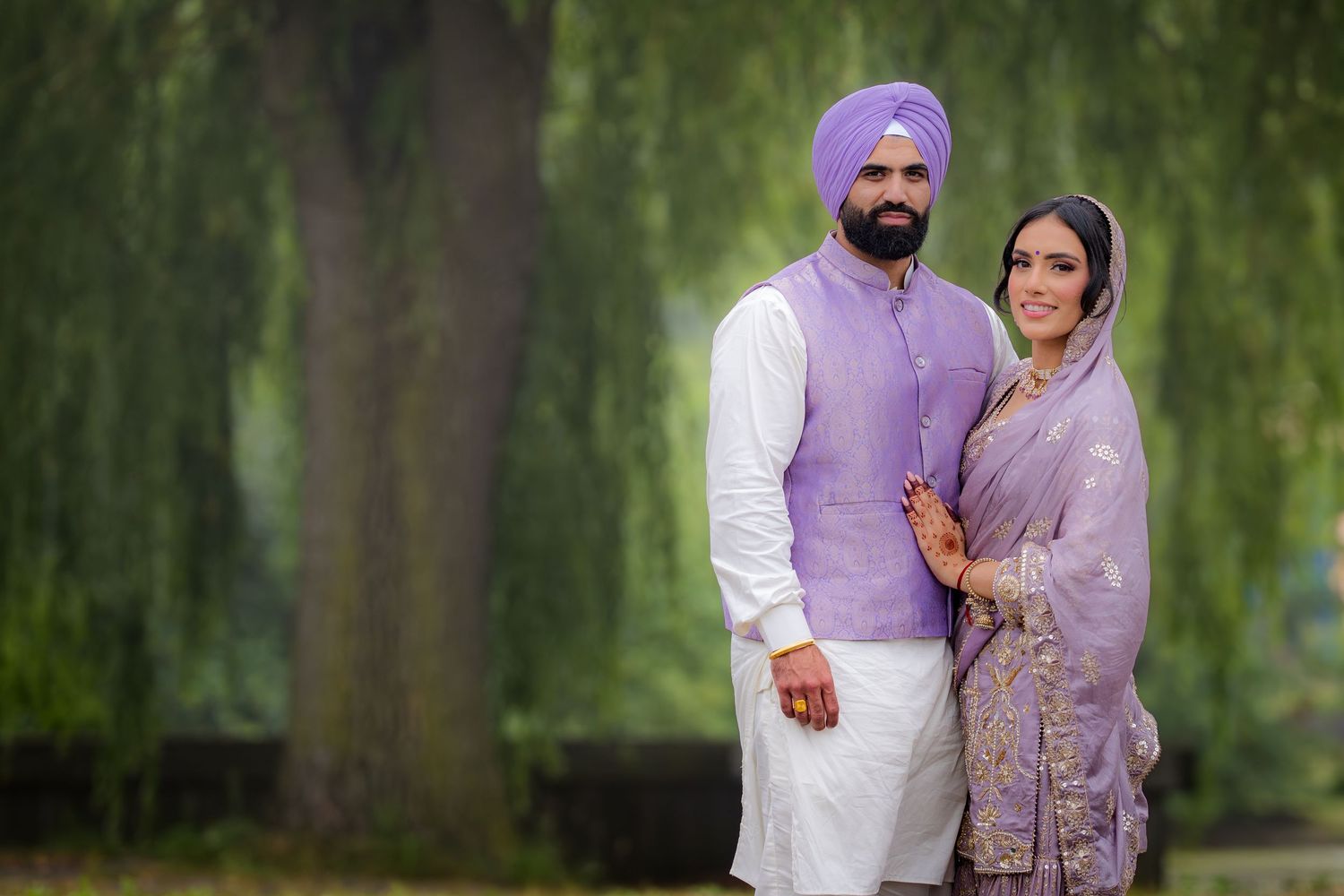 A couple in coordinated purple and white outfits poses together under willow trees in a park setting.