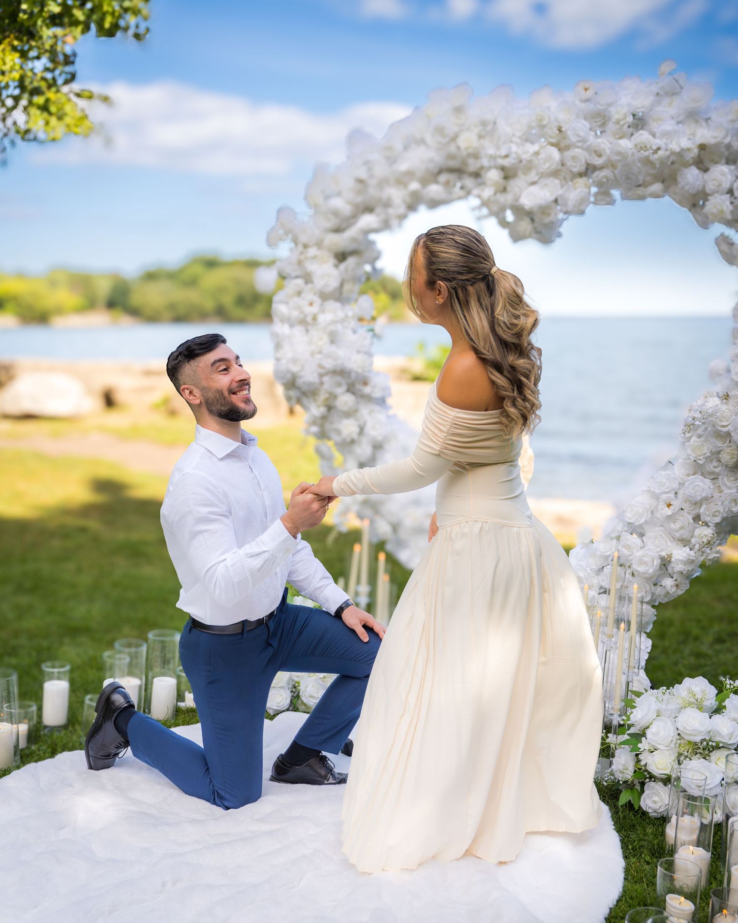 Romantic proposal setup with white floral arch and candles on a grassy oceanfront location during sunset.