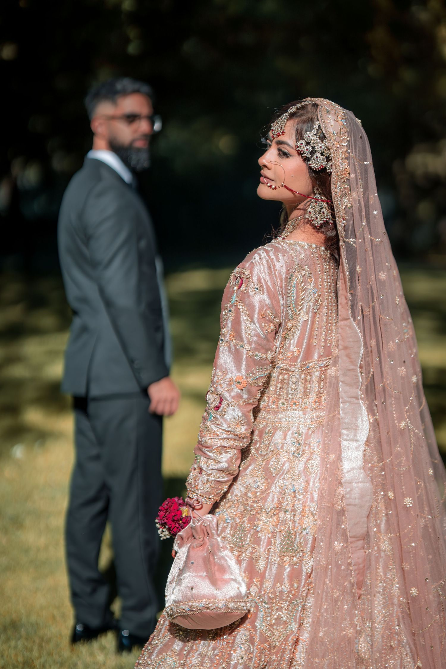 Artistic wedding photo showing bride in peach lehenga with groom in background during outdoor shoot.