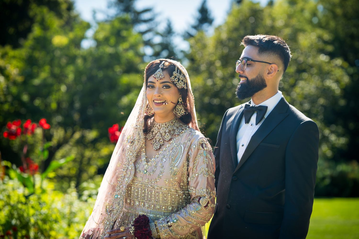 Traditional South Asian wedding couple poses outdoors in elegant attire with natural greenery and sunlight in background.