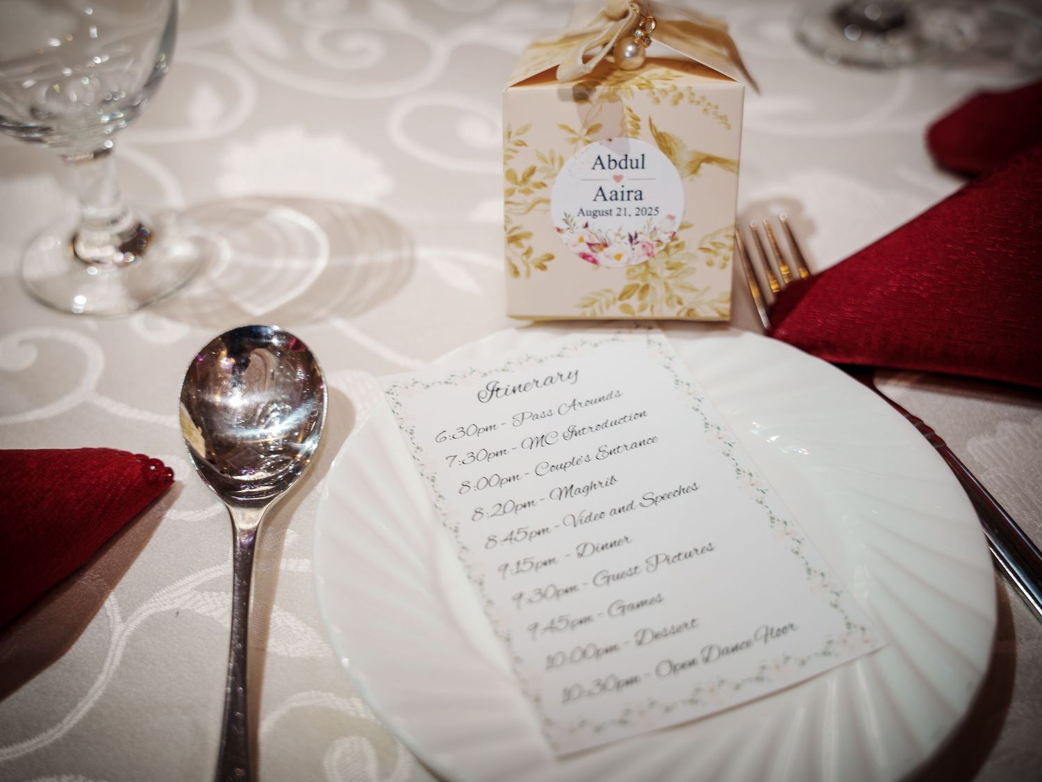 Wedding reception place setting with menu card, favor box, and red napkin on white tablecloth.