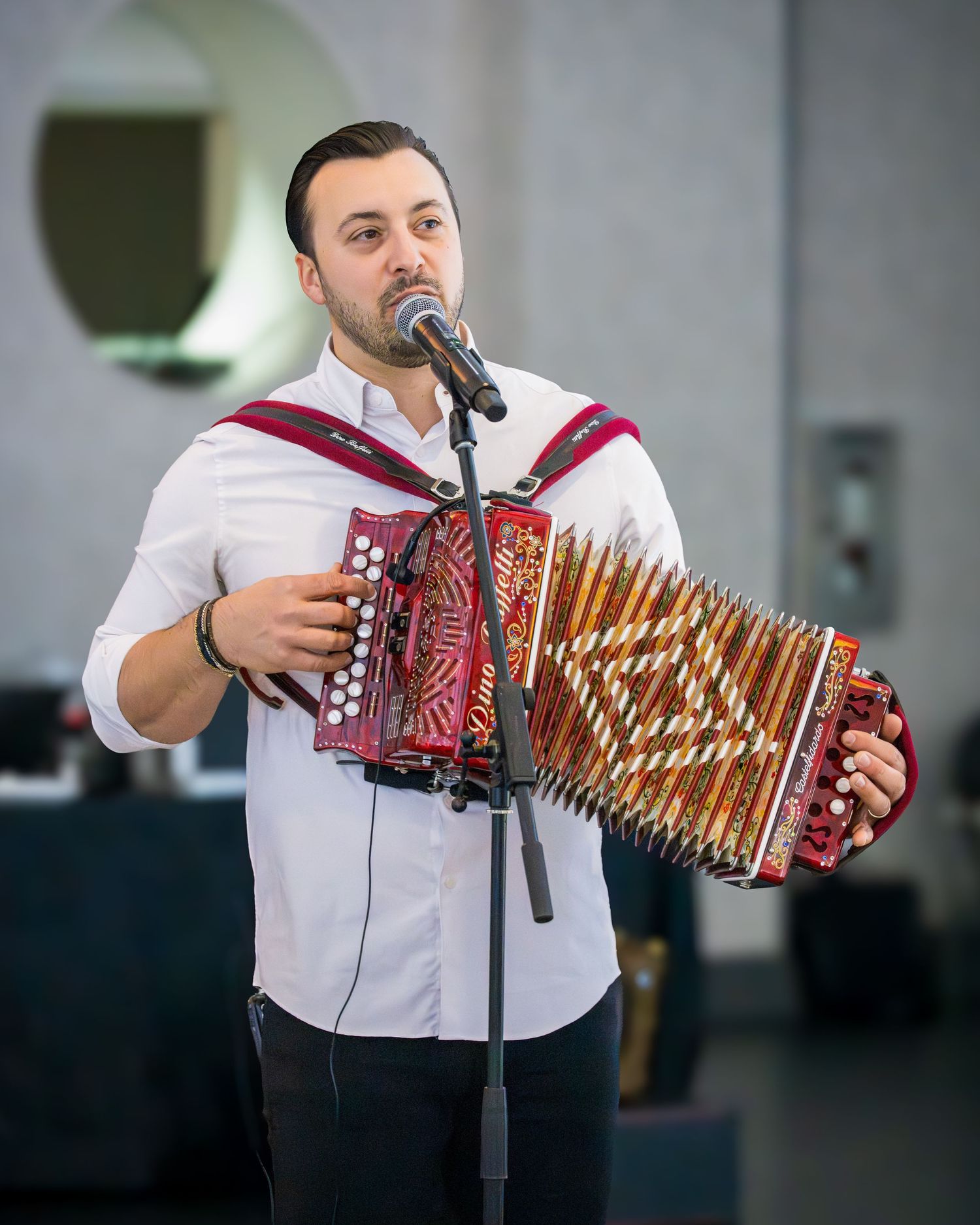 Young male musician playing a red accordion and singing into a microphone at a formal event.