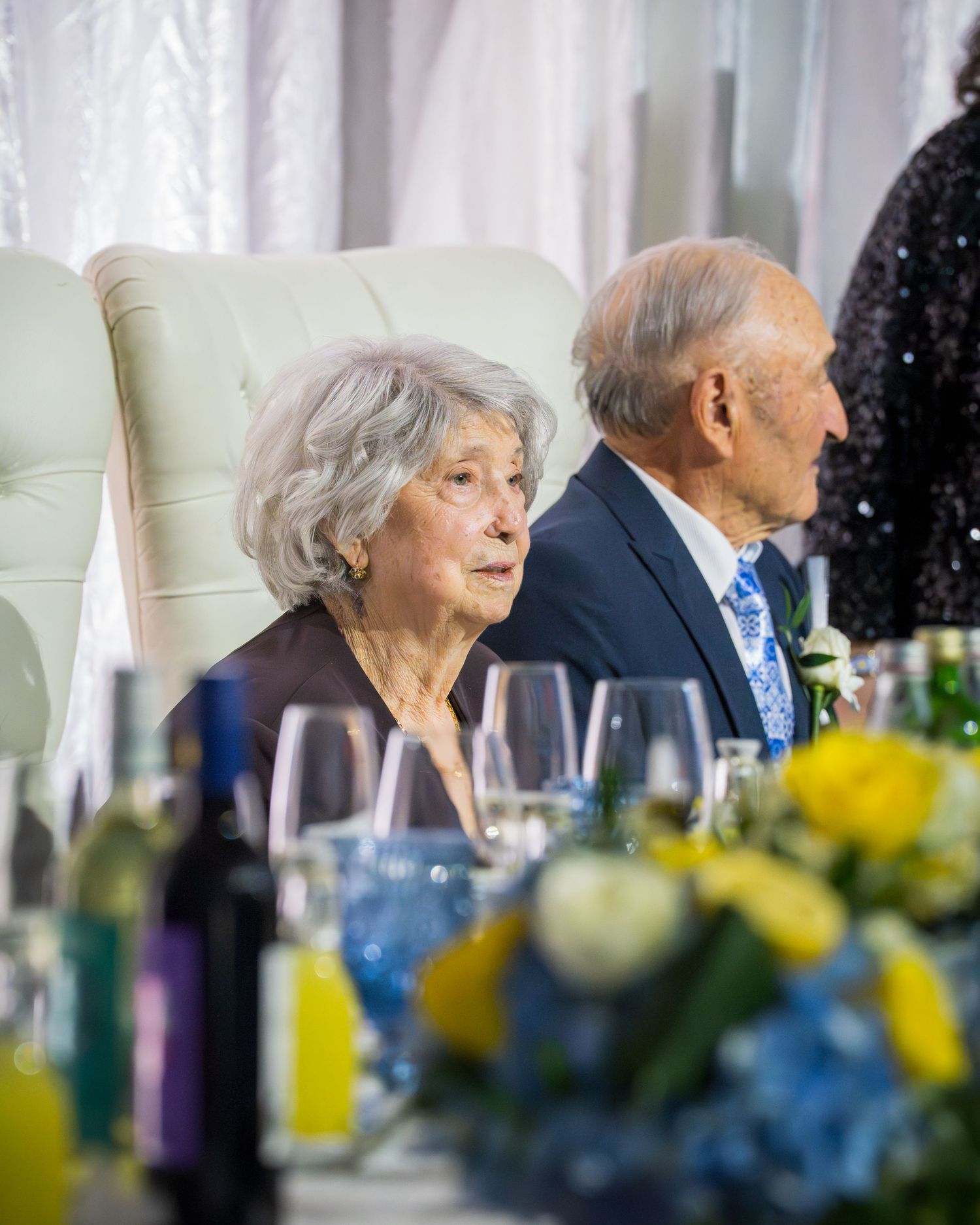 Elderly couple seated at wedding reception head table with yellow and white floral centerpieces.