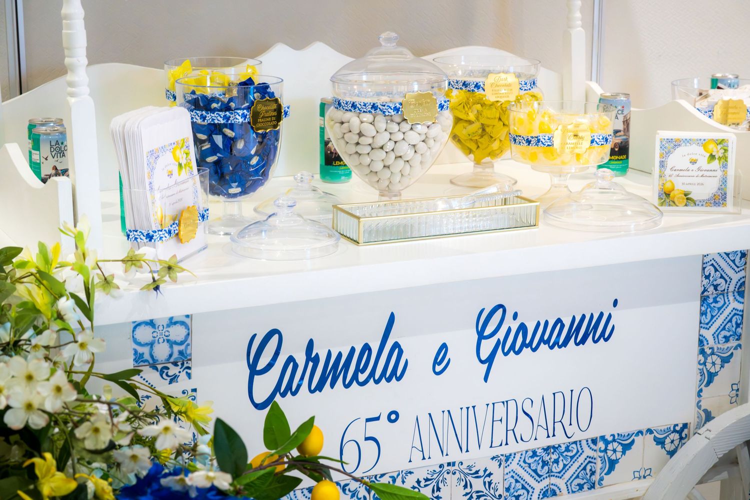 White dessert table labeled Carmela e Giovanni 65 Anniversario with blue and yellow candy jars and flowers.