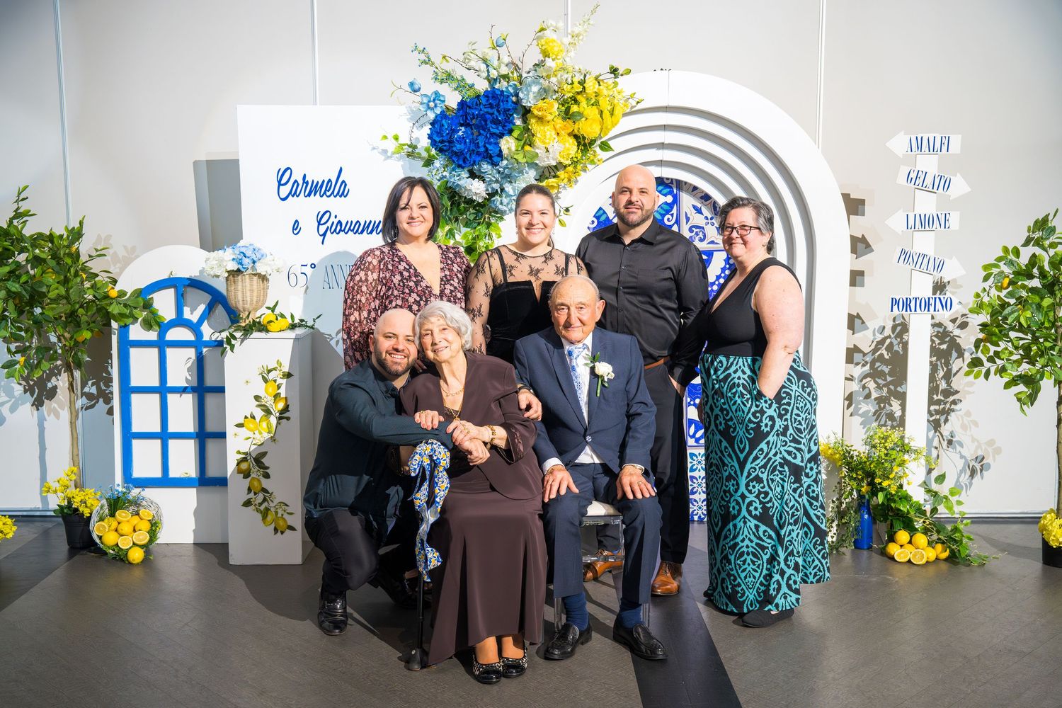 Family group photo at Carmela and Giovanni 65th anniversary party in front of decorative Italian-themed arch backdrop.