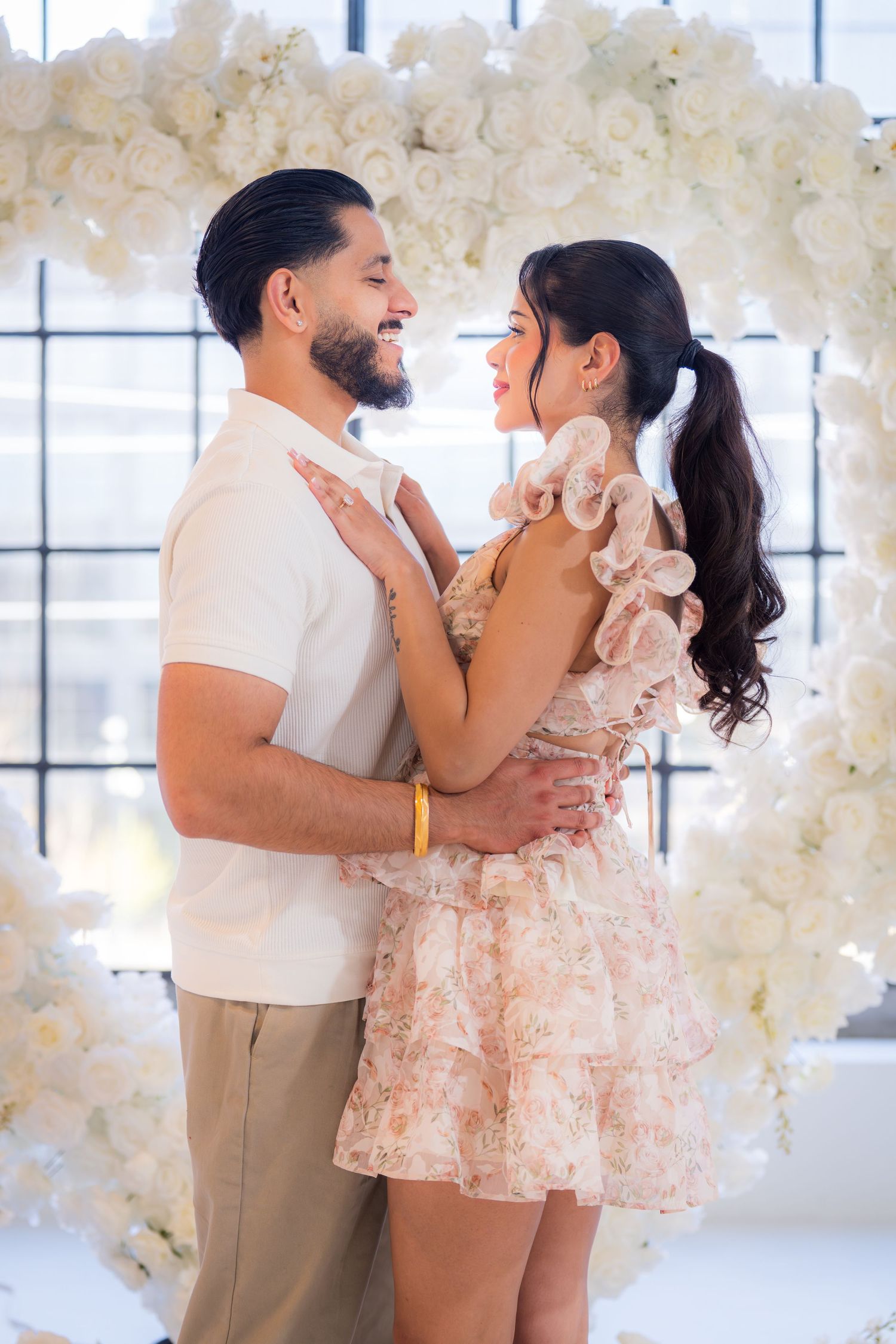 Happy couple embracing and smiling beneath white floral heart arch backdrop.