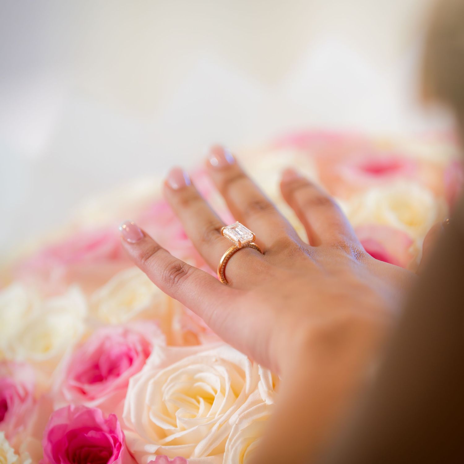 Close-up of elegant emerald-cut engagement ring on hand resting over pink and white roses.