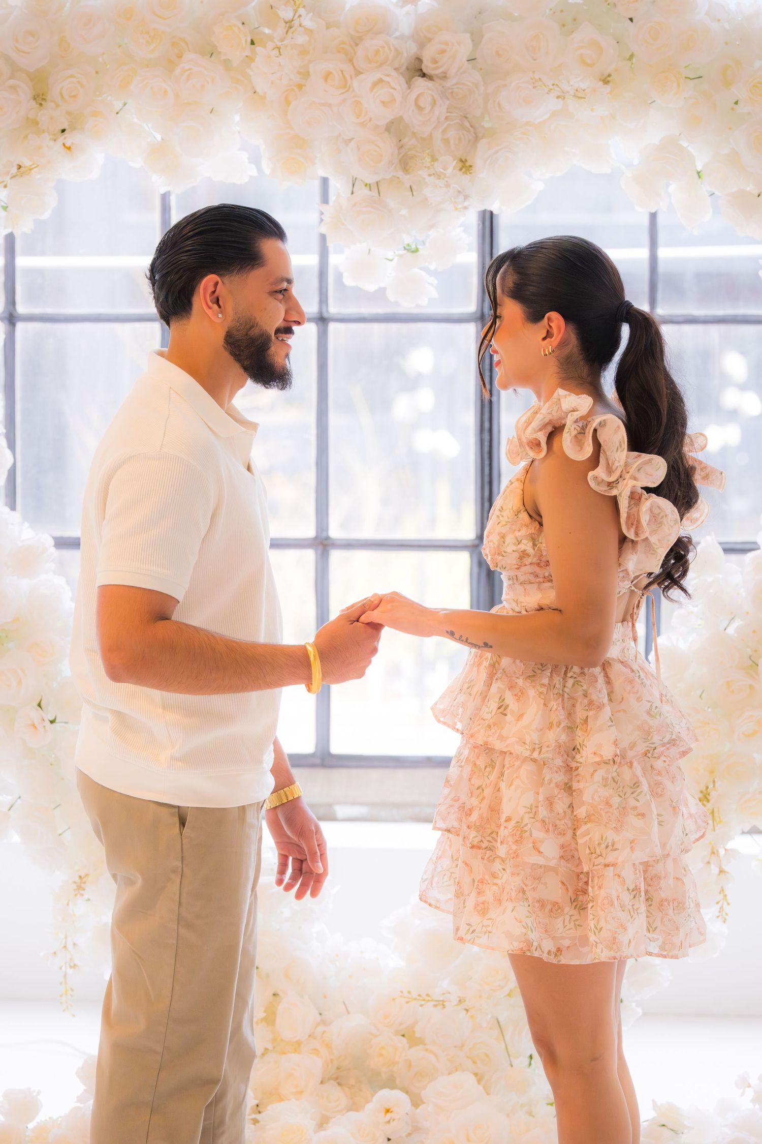 Man and woman holding hands and smiling at each other under an elegant white floral arch.