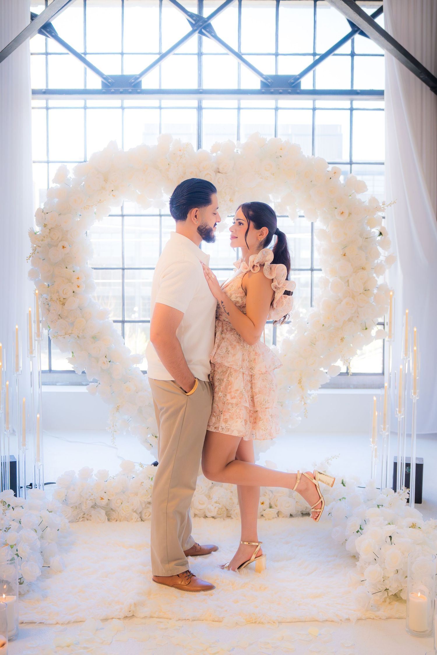 Couple embracing in front of large white floral heart arch with candles and fairy lights.