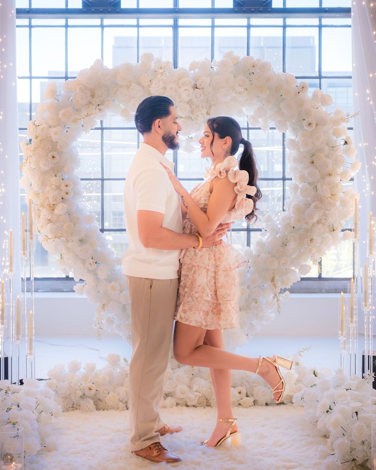Couple embracing and smiling in front of large white floral heart arch with lights.