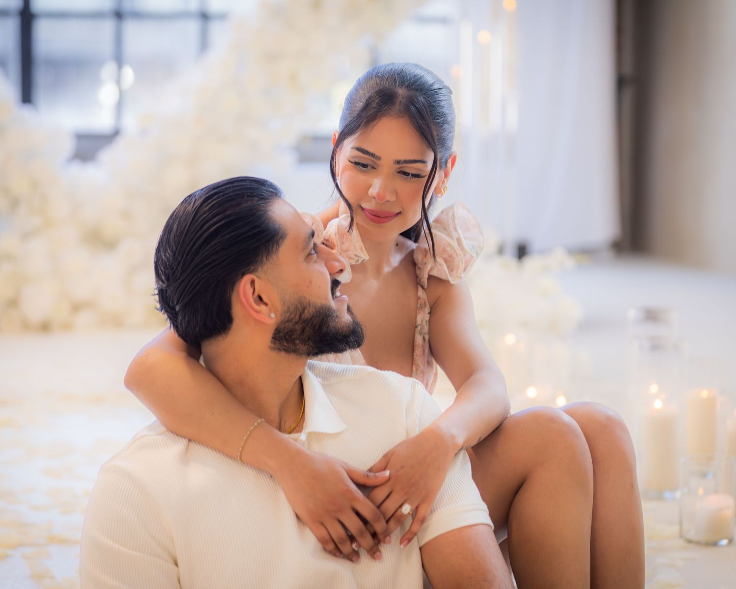 An engaged couple shares a tender intimate moment surrounded by candles and white rose petals.