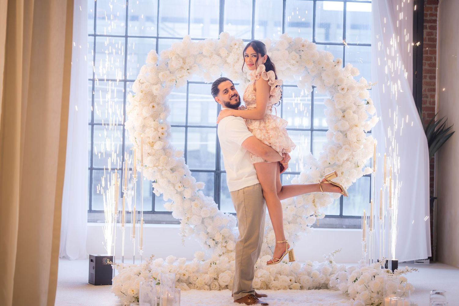 Couple celebrating joyfully in front of a large white floral heart arch with sparklers.