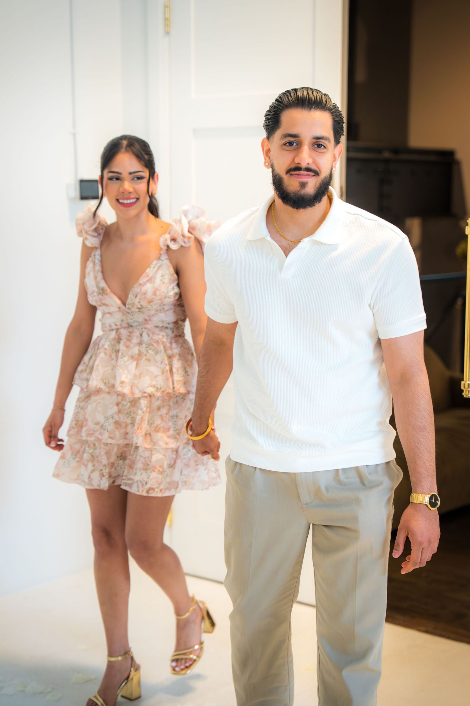 Smiling couple holding hands walking through doorway, man in white polo and woman in floral dress.