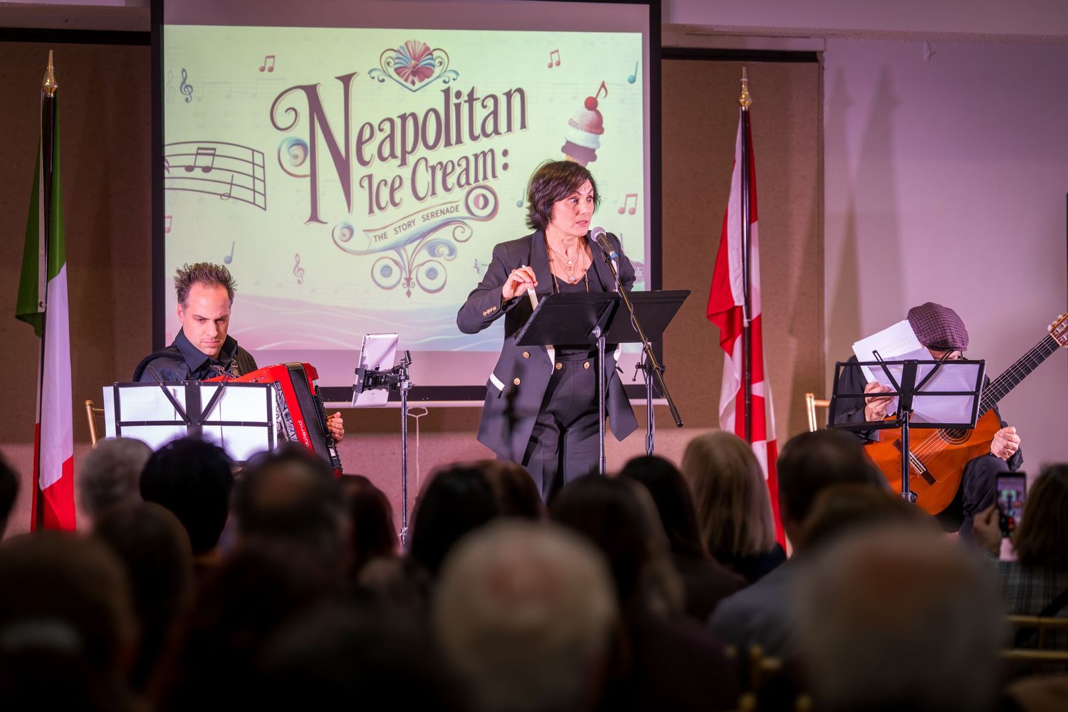 Three performers on stage at a Neapolitan Ice Cream concert with Italian flags and audience in foreground.