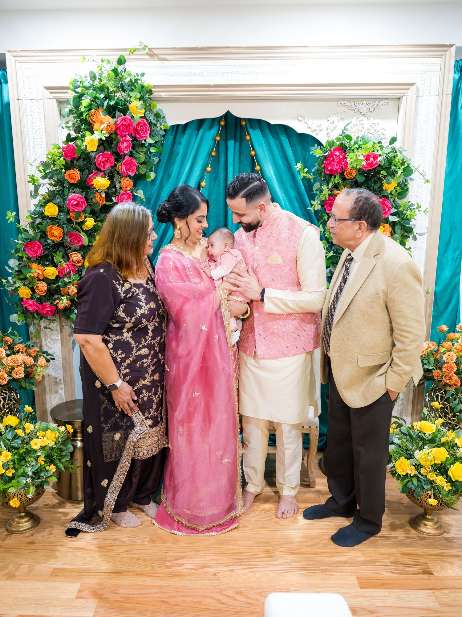 Family gathering around newborn baby in pink outfit at decorated Lohri celebration with teal floral arch.