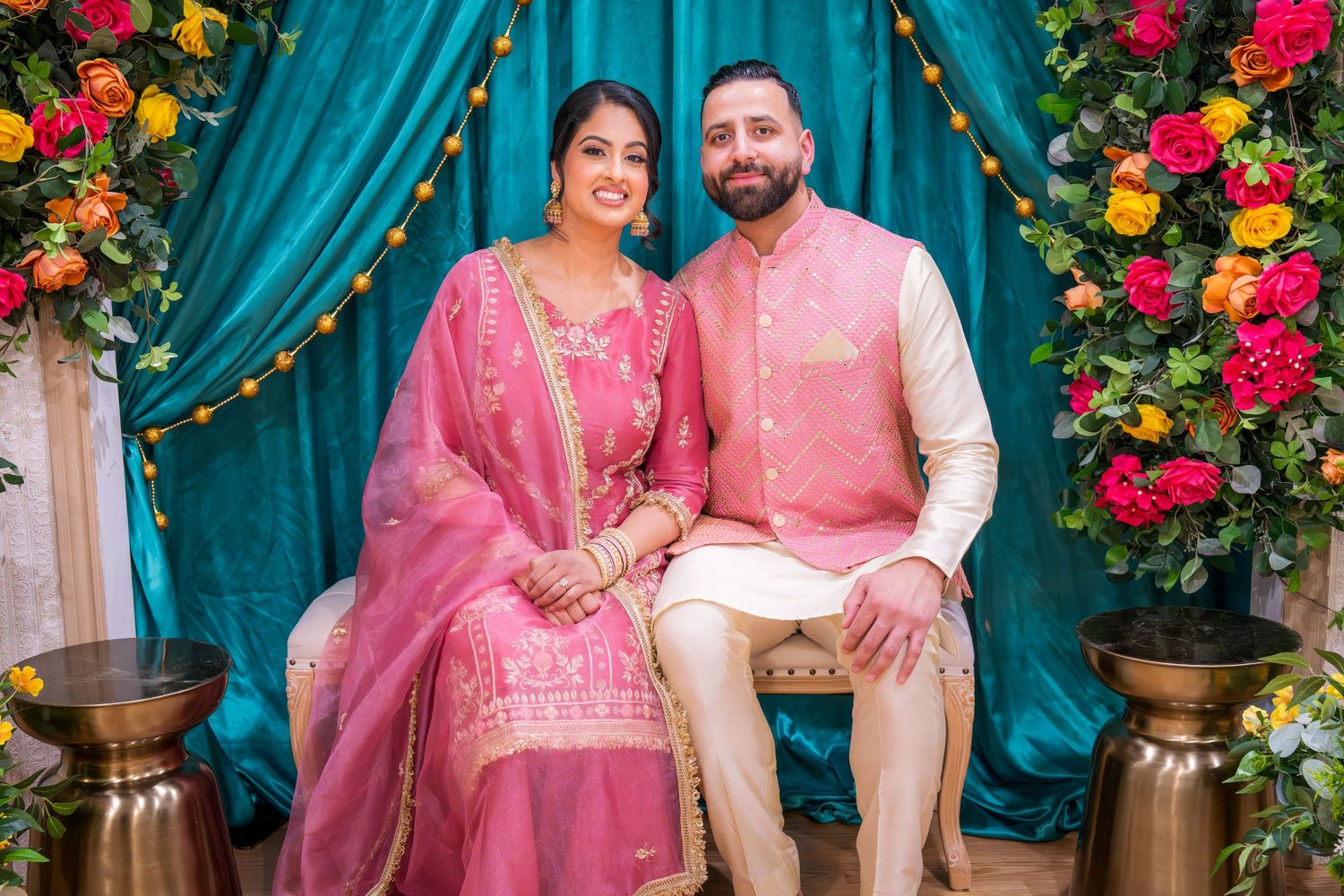 South Asian couple in coordinating pink traditional outfits seated at floral decorated celebration.