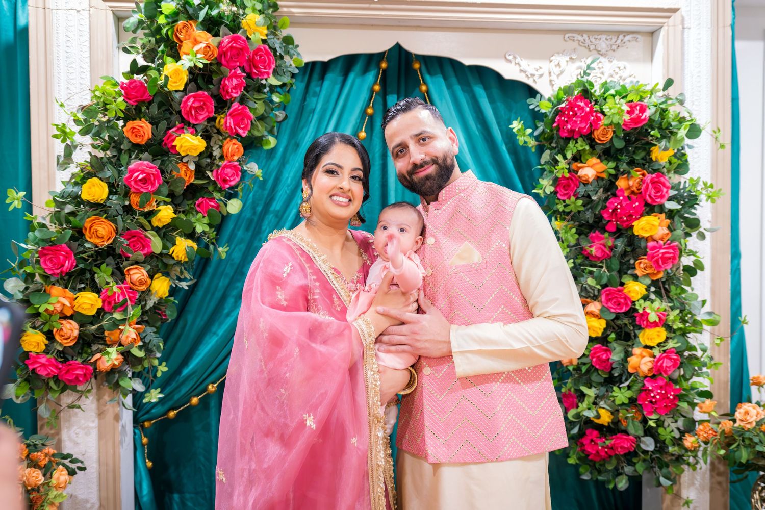 A smiling couple in matching pink traditional Indian outfits holding their baby at a vibrant floral ceremony.