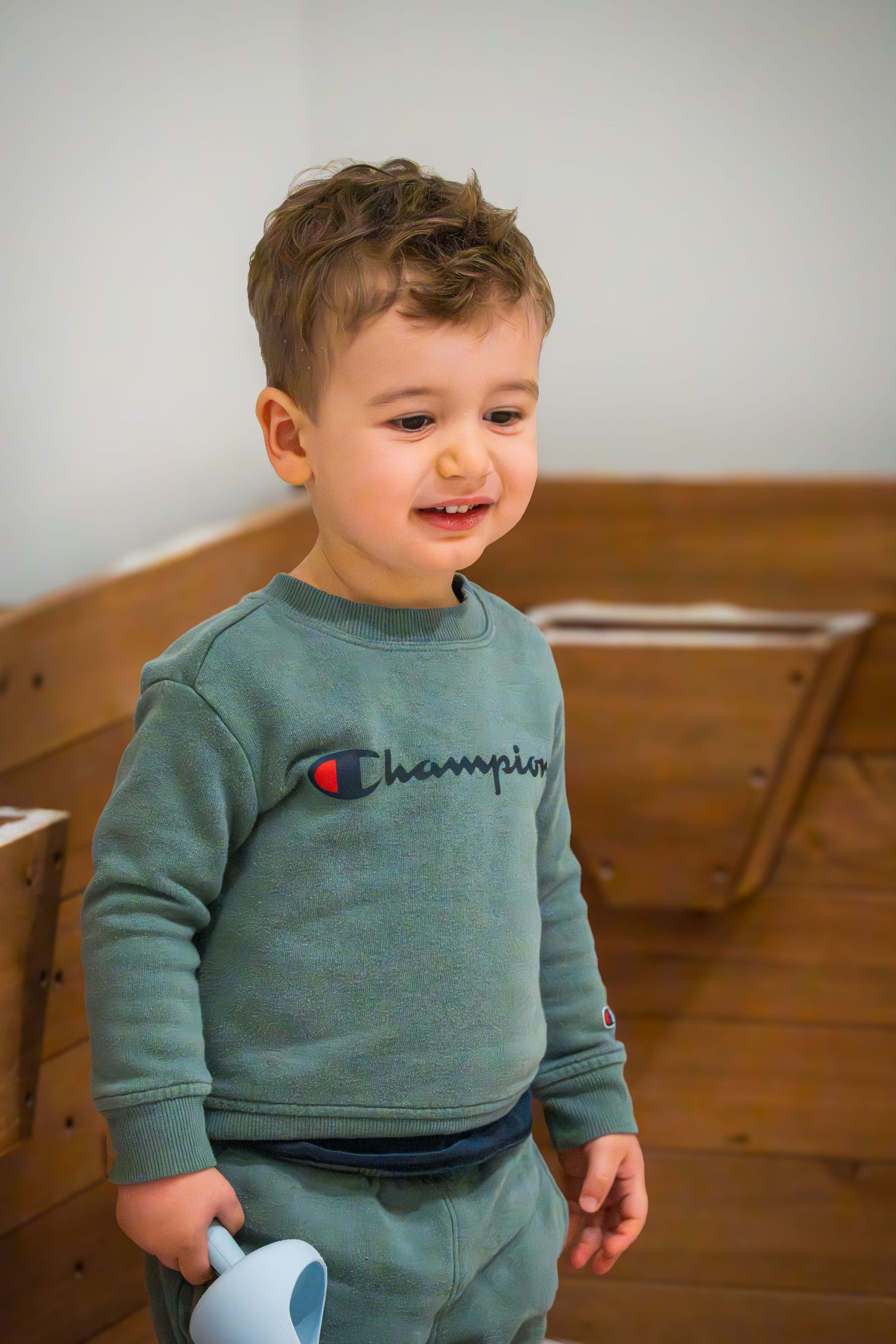 Happy toddler boy in a green Champion crewneck sweatshirt holding a light blue cup indoors.