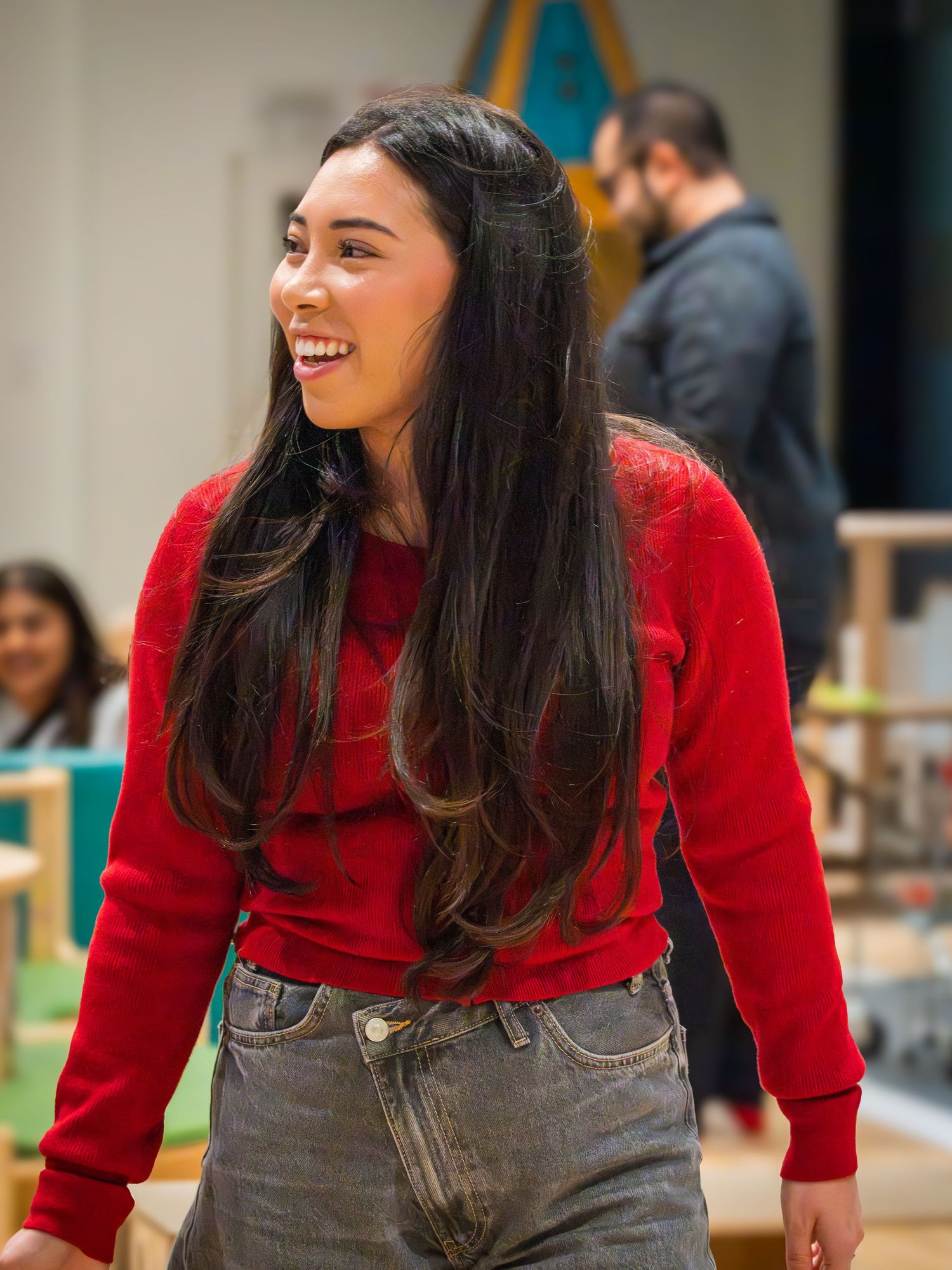 A young woman with long dark hair laughs while wearing a red sweater and gray jeans indoors.