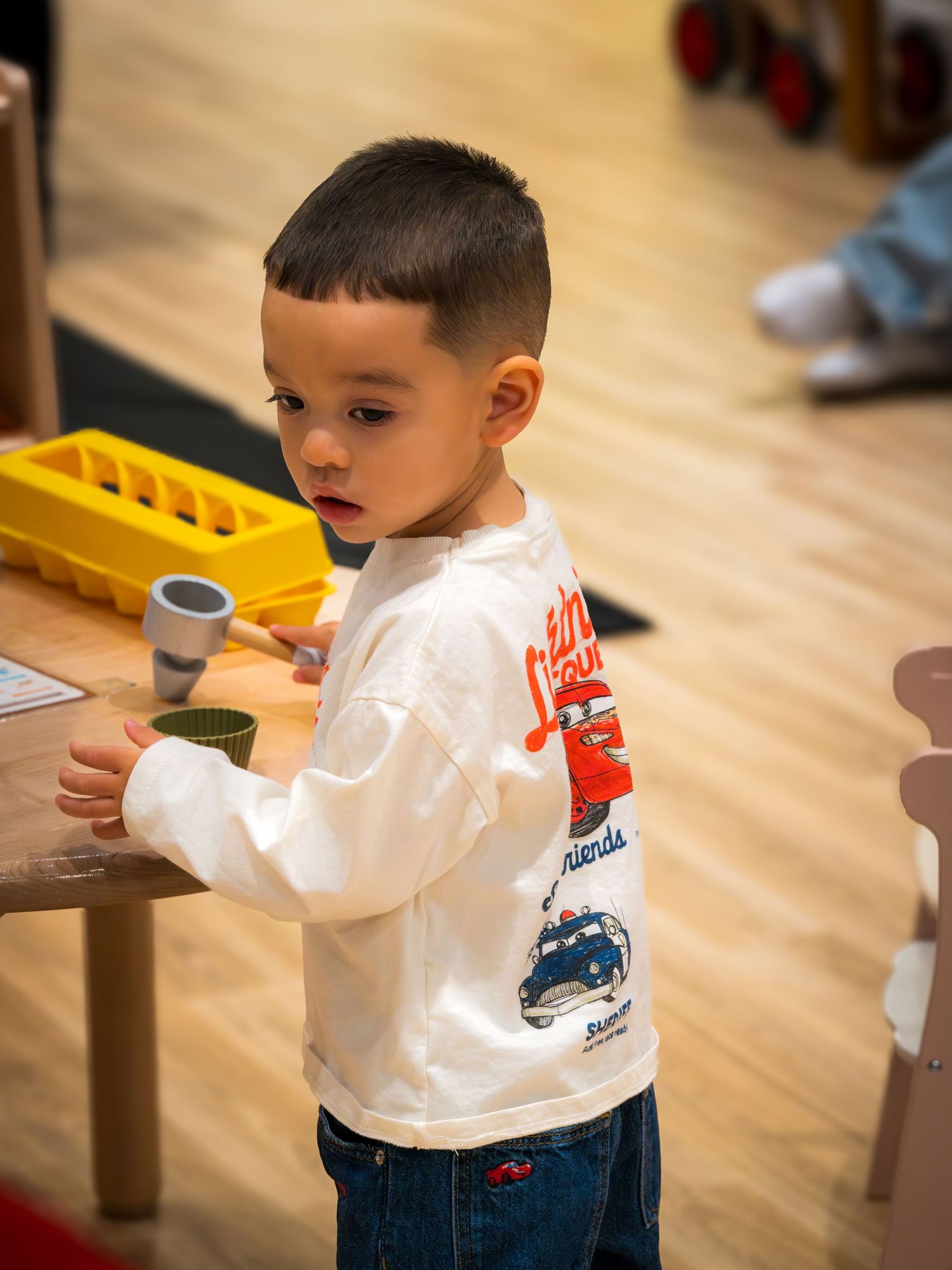 Young boy in white graphic sweatshirt and jeans playing with toys at a wooden table.