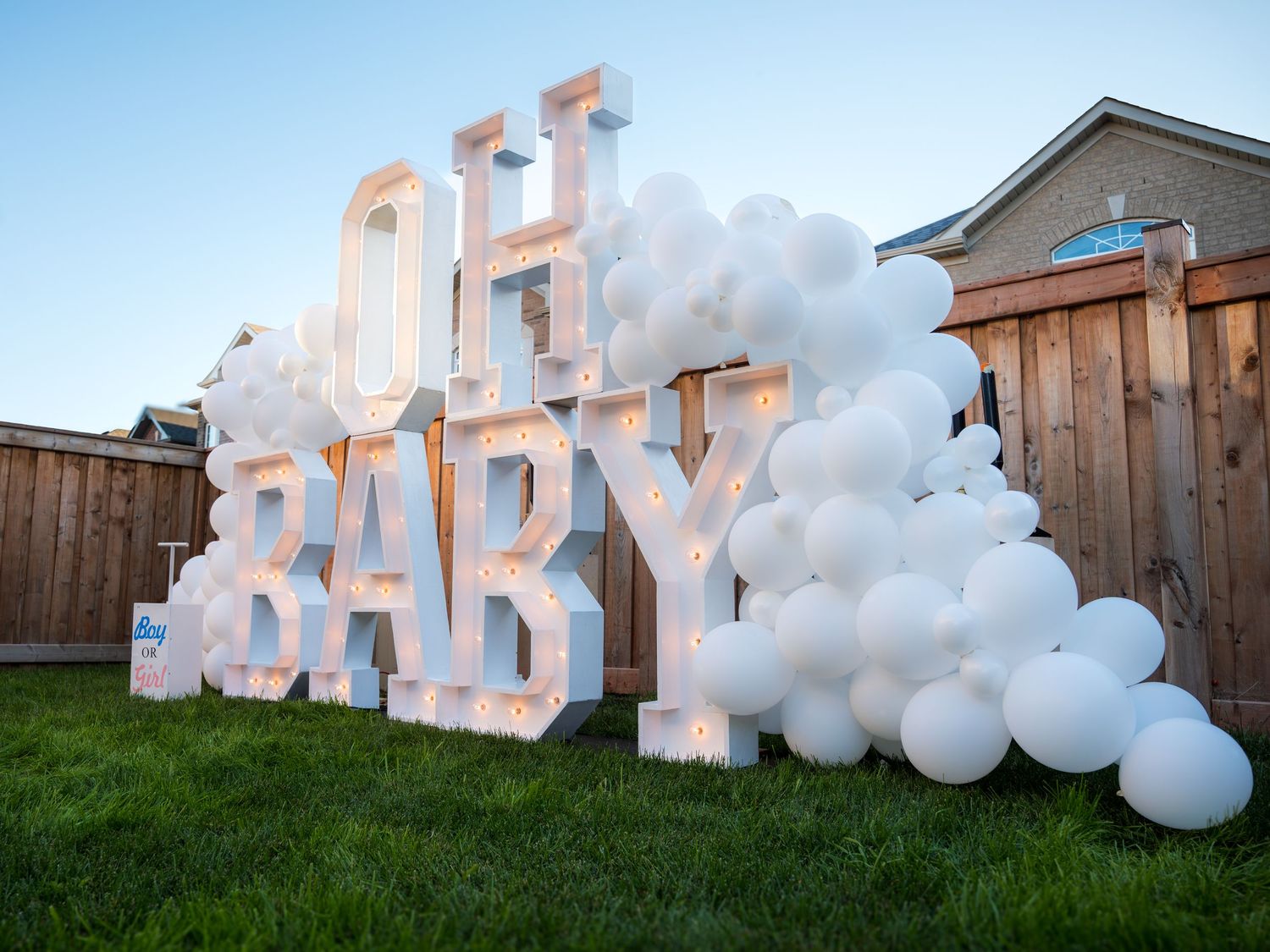 Illuminated OH BABY marquee letters surrounded by white balloons at an outdoor baby shower.