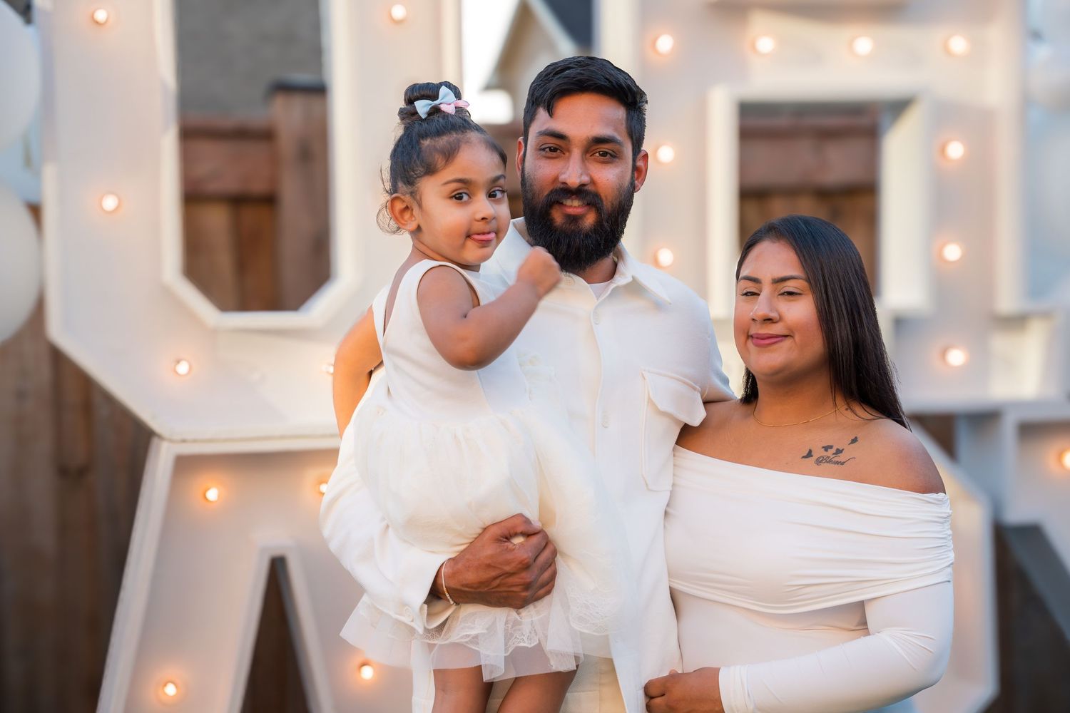 Smiling couple in white outfits holding young daughter at outdoor baby shower celebration with marquee letters backdrop.