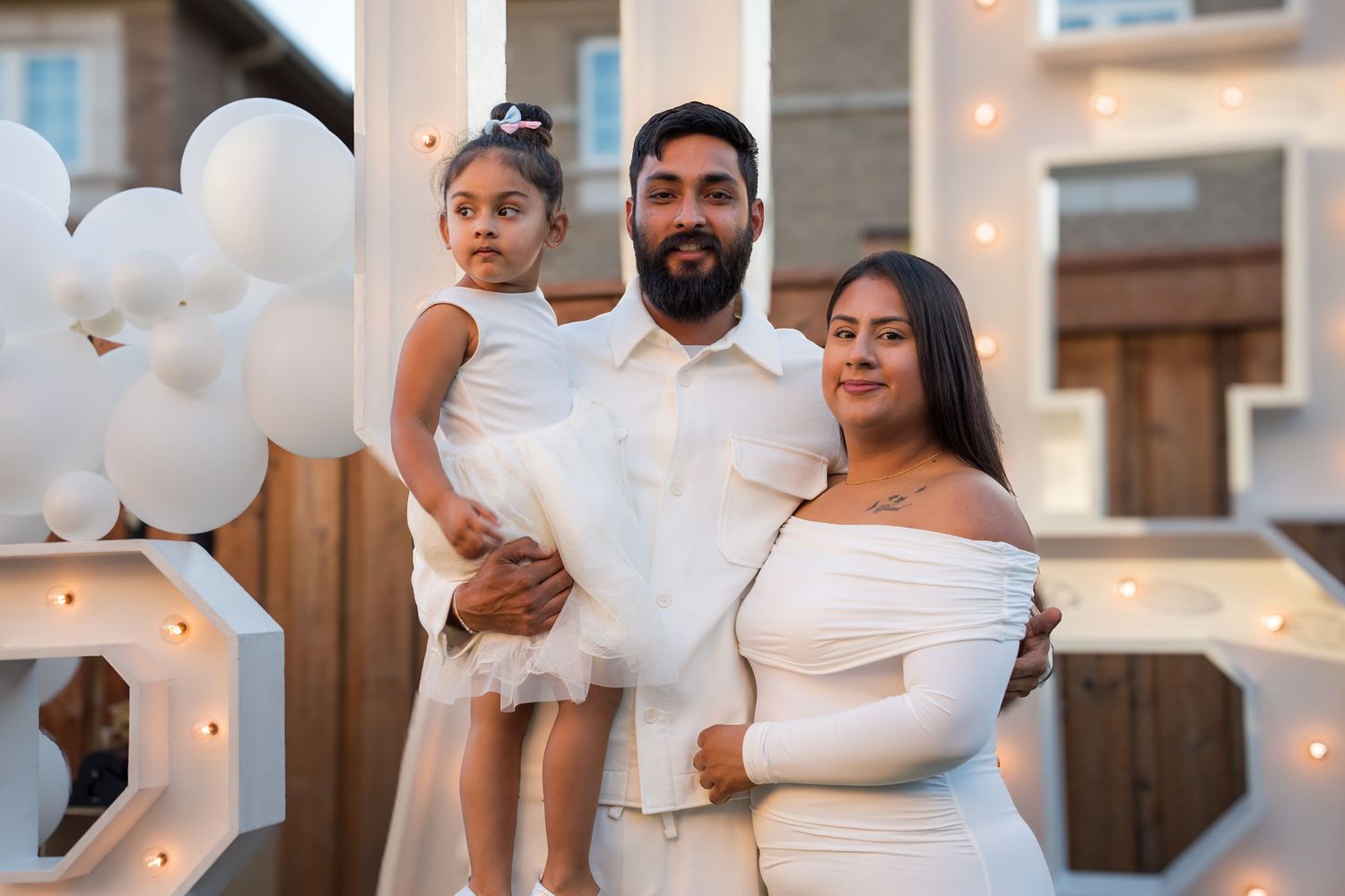 A family of three in white outfits poses outdoors with white balloons and light-up letters at a celebration.