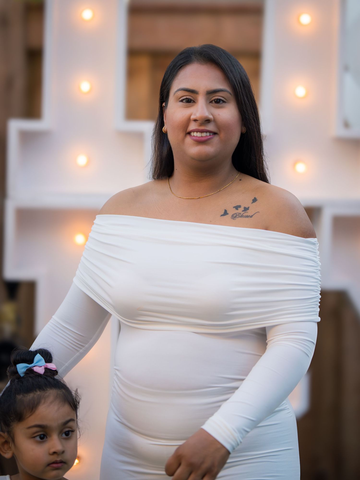 Pregnant woman in white off-shoulder maternity dress posing with young daughter at baby shower.