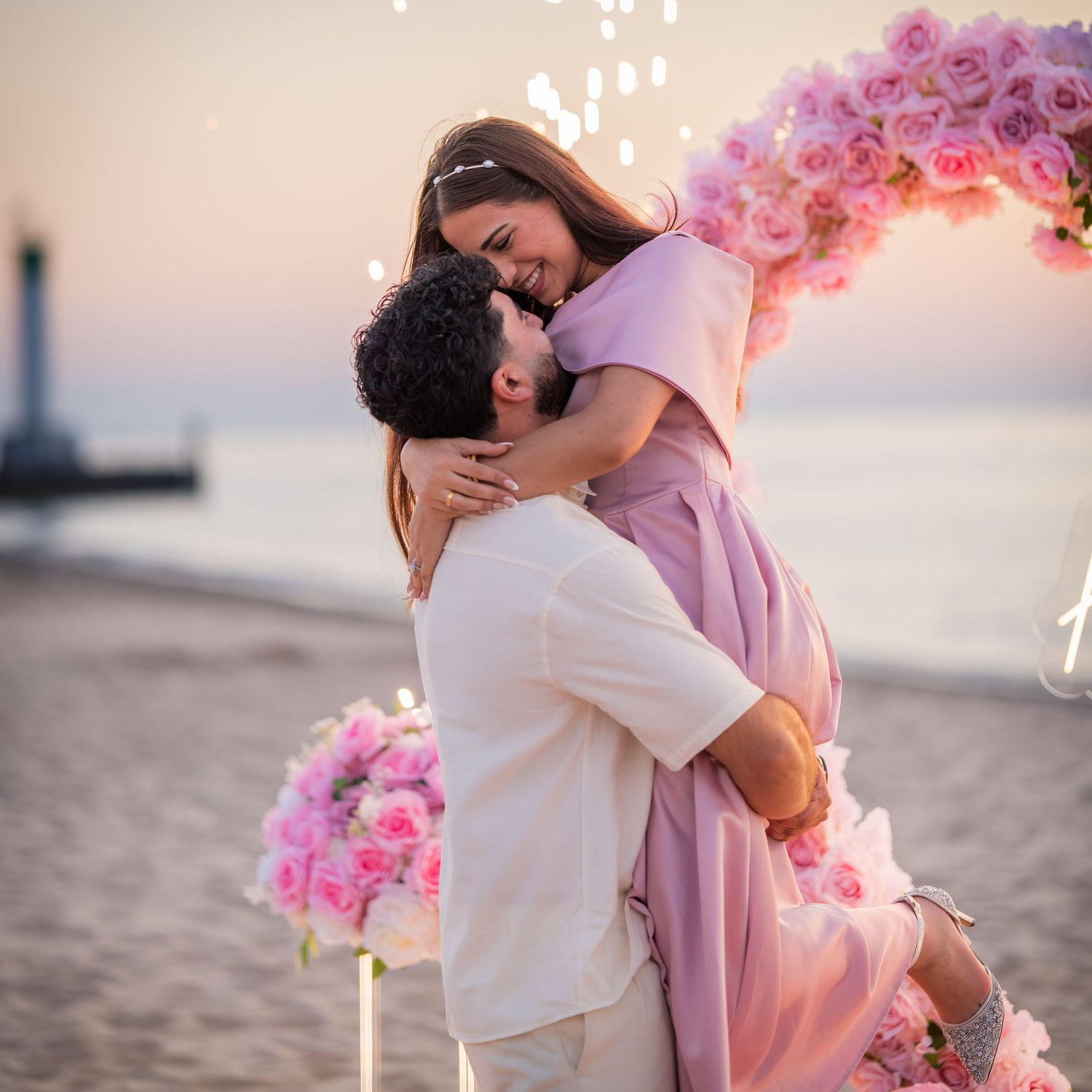 A romantic beach proposal setup with a couple embracing under a pink floral arch at sunset.