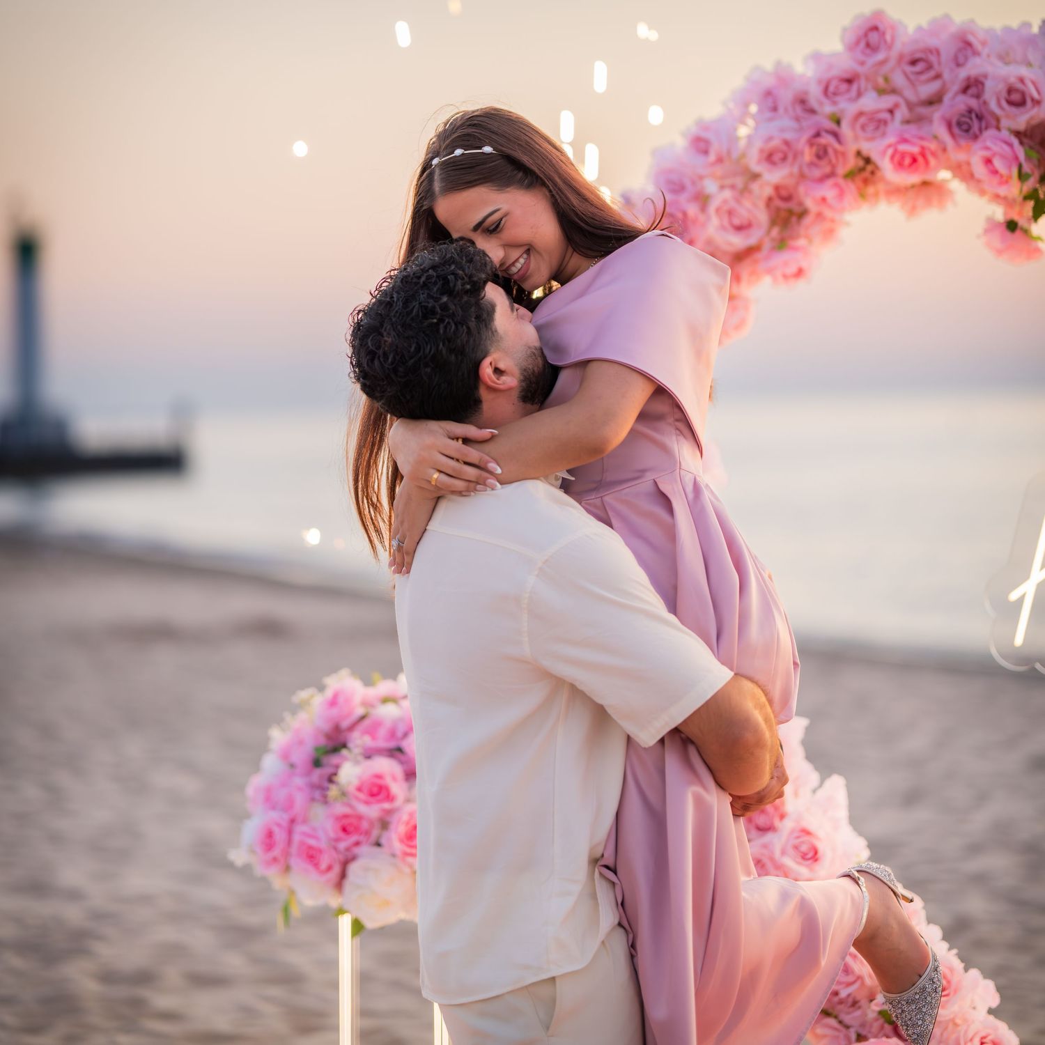 An intimate beach moment captured with a pink floral arch and lighthouse silhouette at dusk.