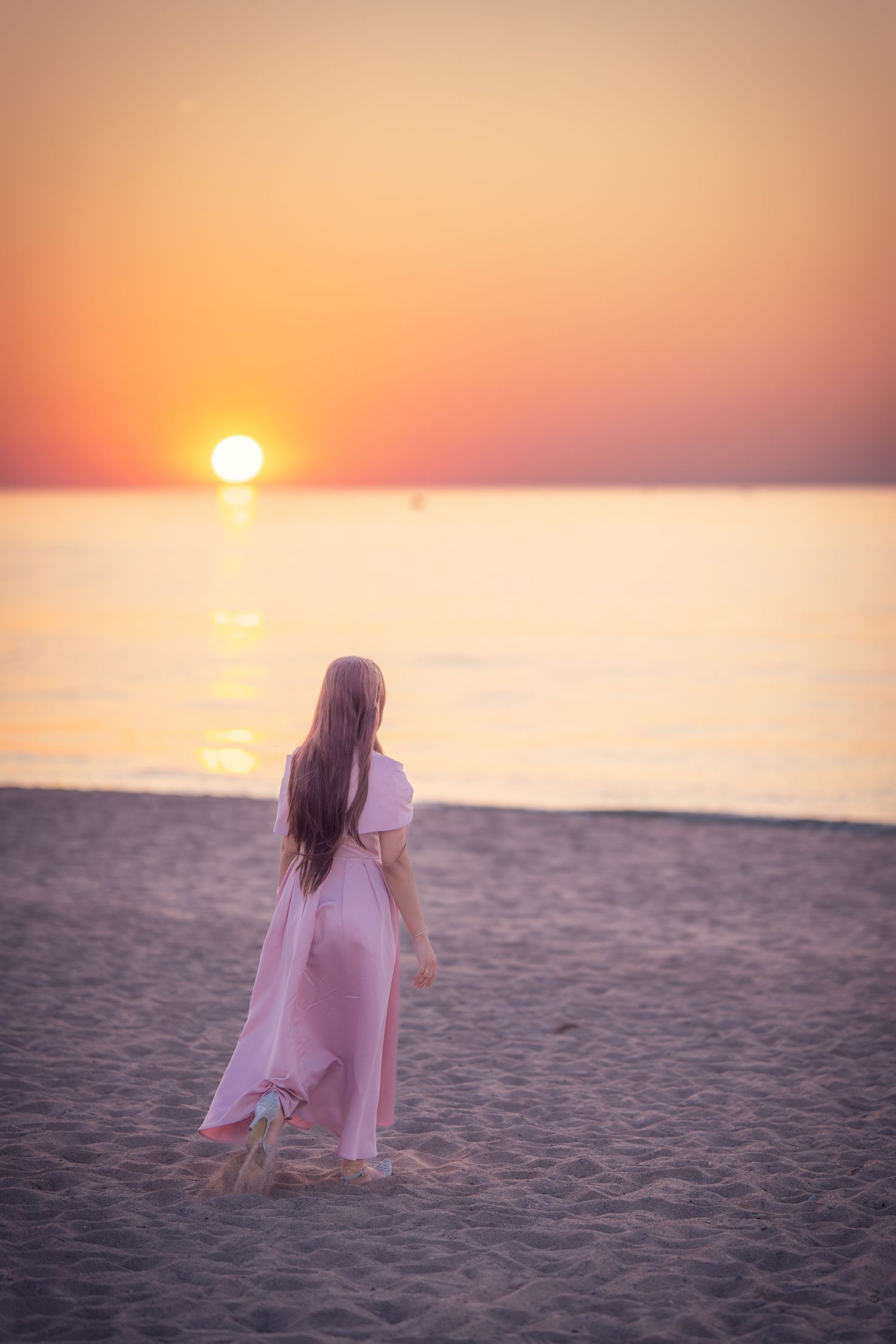 A person in a pink dress stands on a beach during sunset with the sun reflecting on calm waters.