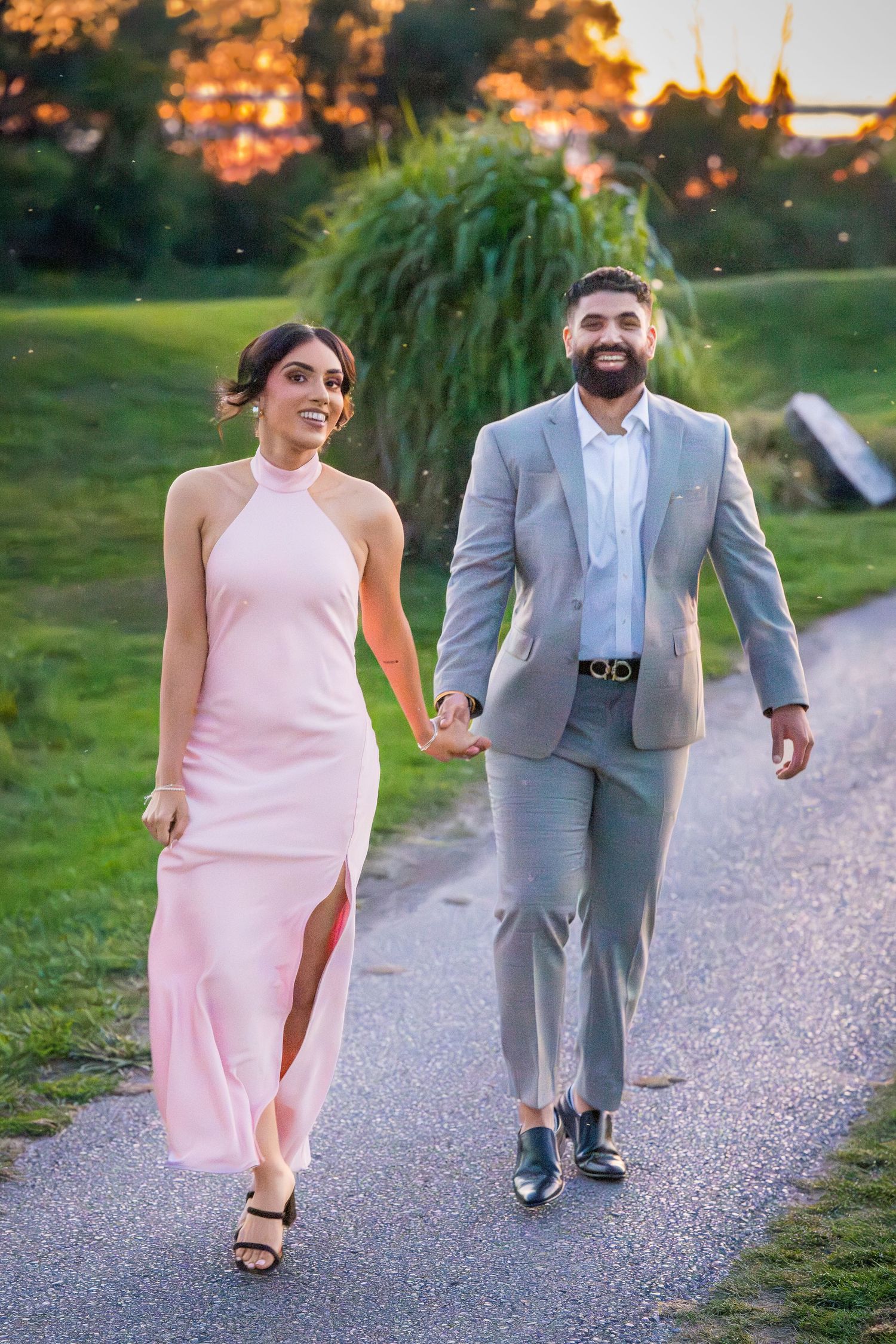 Couple walks along gravel path at sunset wearing pink dress and grey suit.