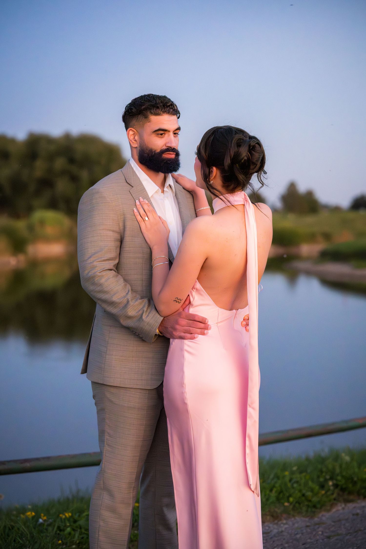 A romantic lakeside portrait of a couple embracing, with one in pink and one in tan formal wear.