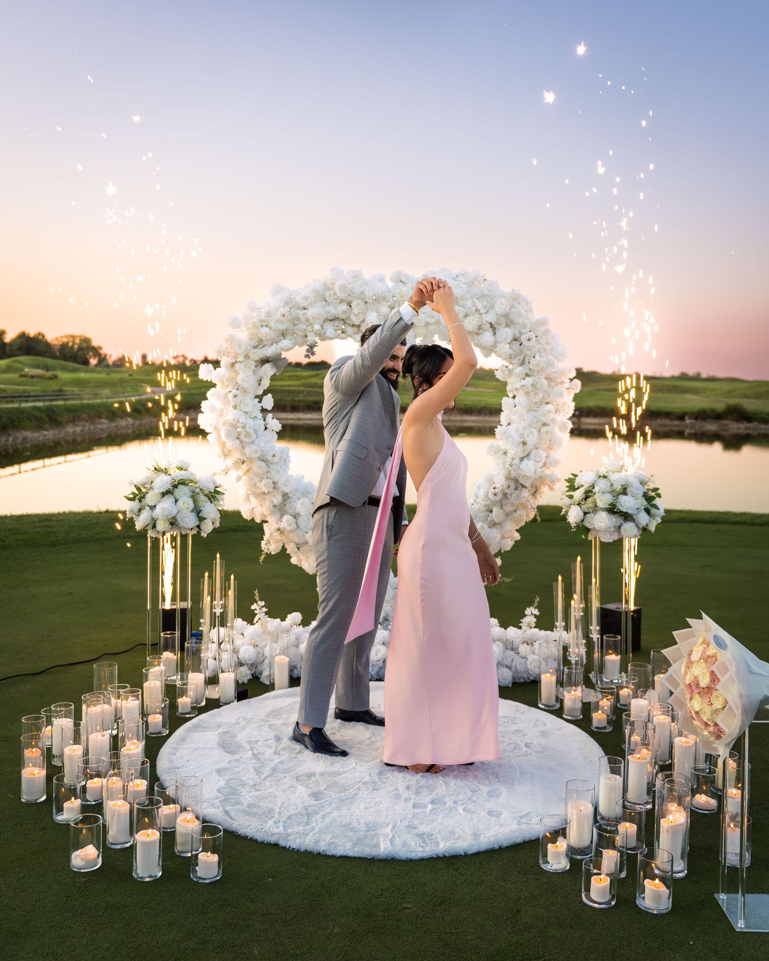 A romantic outdoor wedding ceremony setup with a white floral arch and glowing candle lanterns on a golf course at sunset.