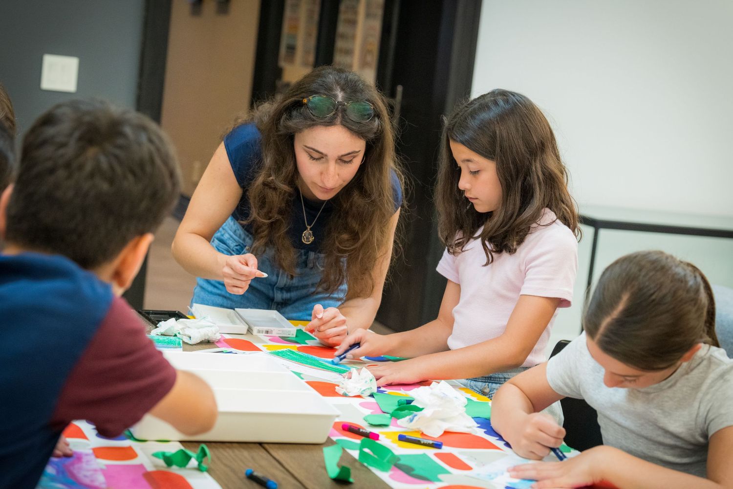 Art teacher assists group of children with colorful paper collage project at a classroom table.