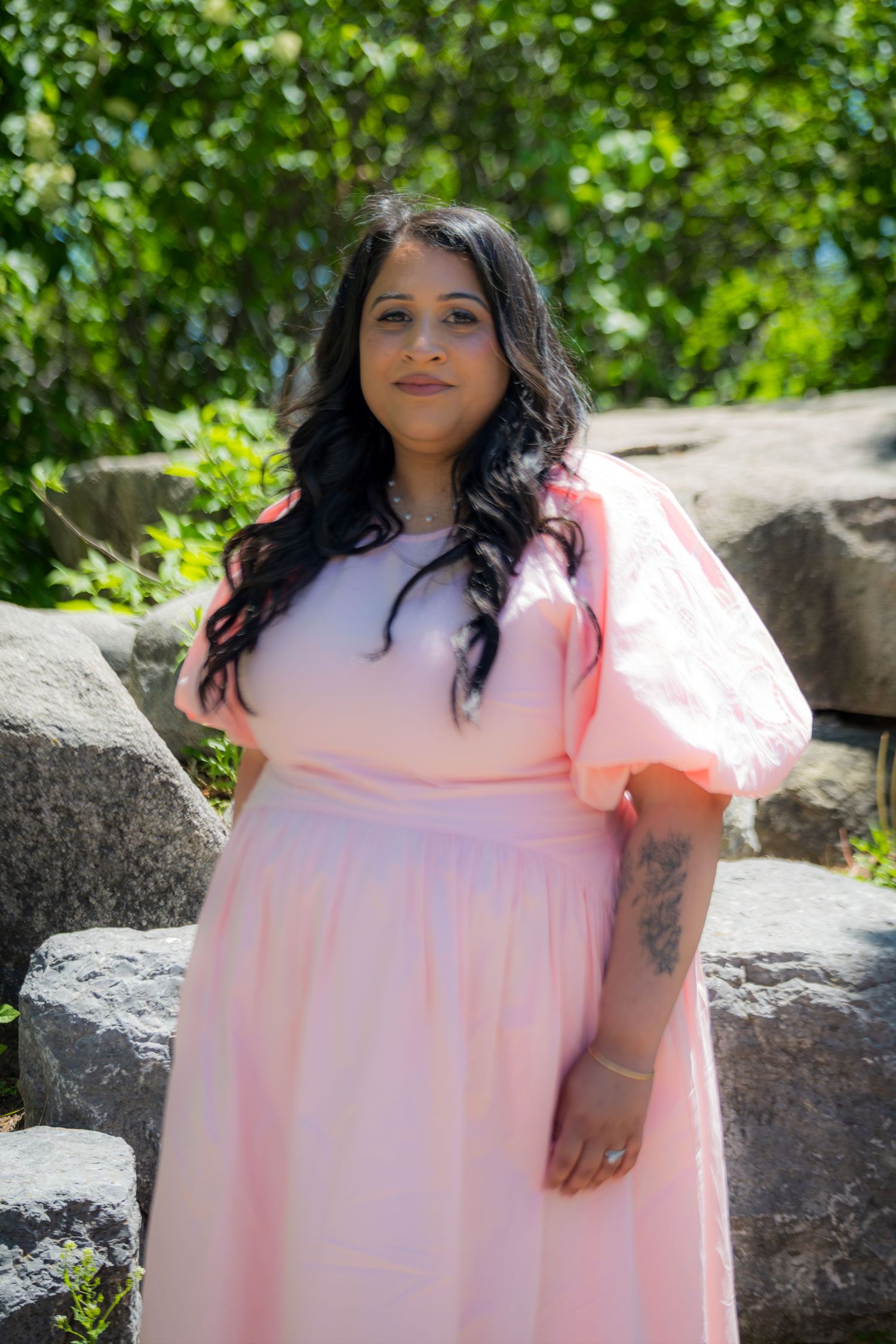 A woman with dark wavy hair wearing a pink puff-sleeve dress stands near rocks in a garden.