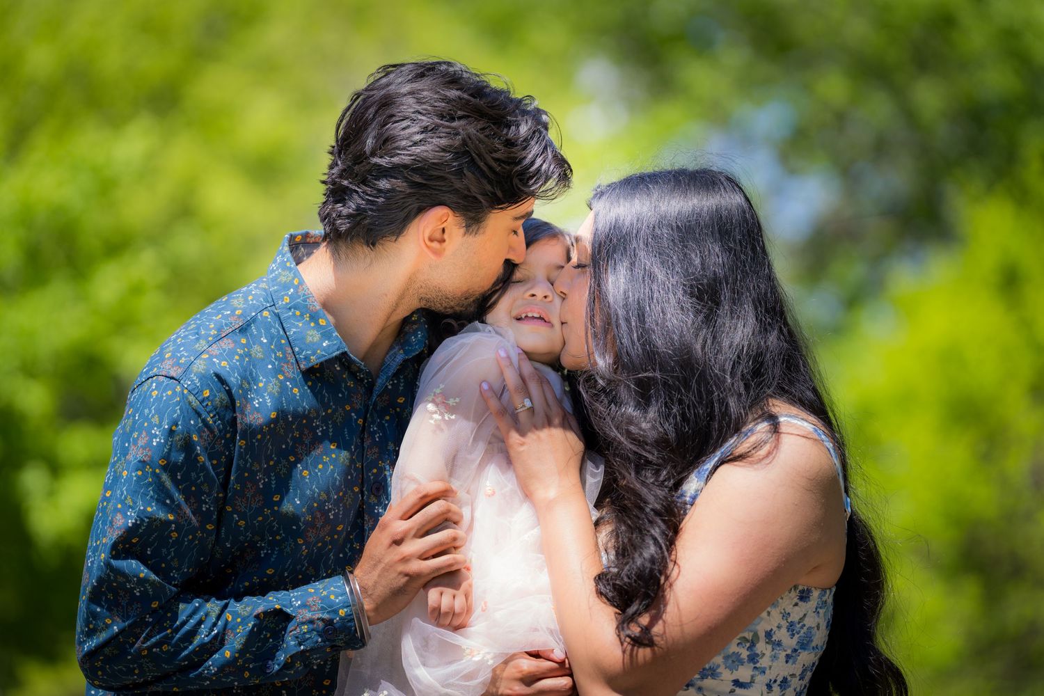 Couple tenderly kisses baby between them in sunny green outdoor setting.