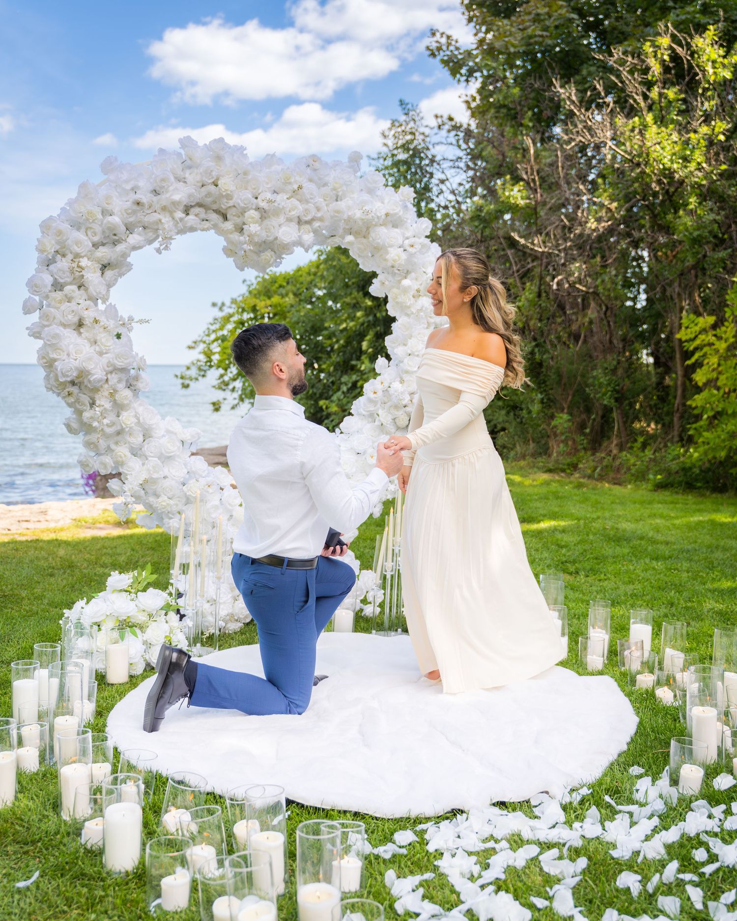 Beautiful proposal moment under a white floral arch surrounded by glowing candles at sunset.