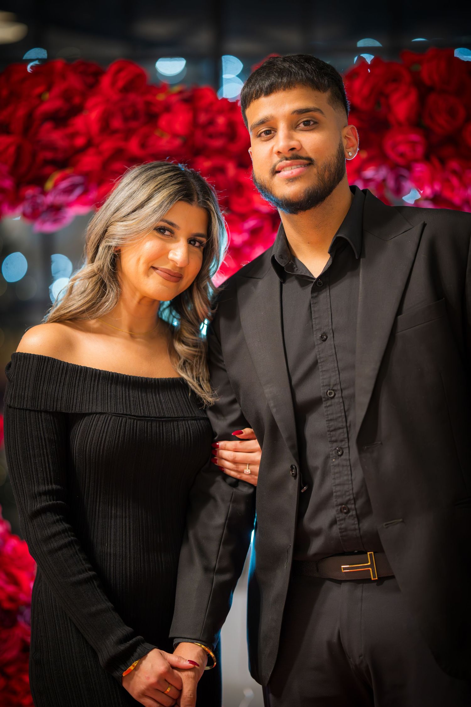 Elegant couple poses against vibrant red rose wall backdrop in formal black attire.